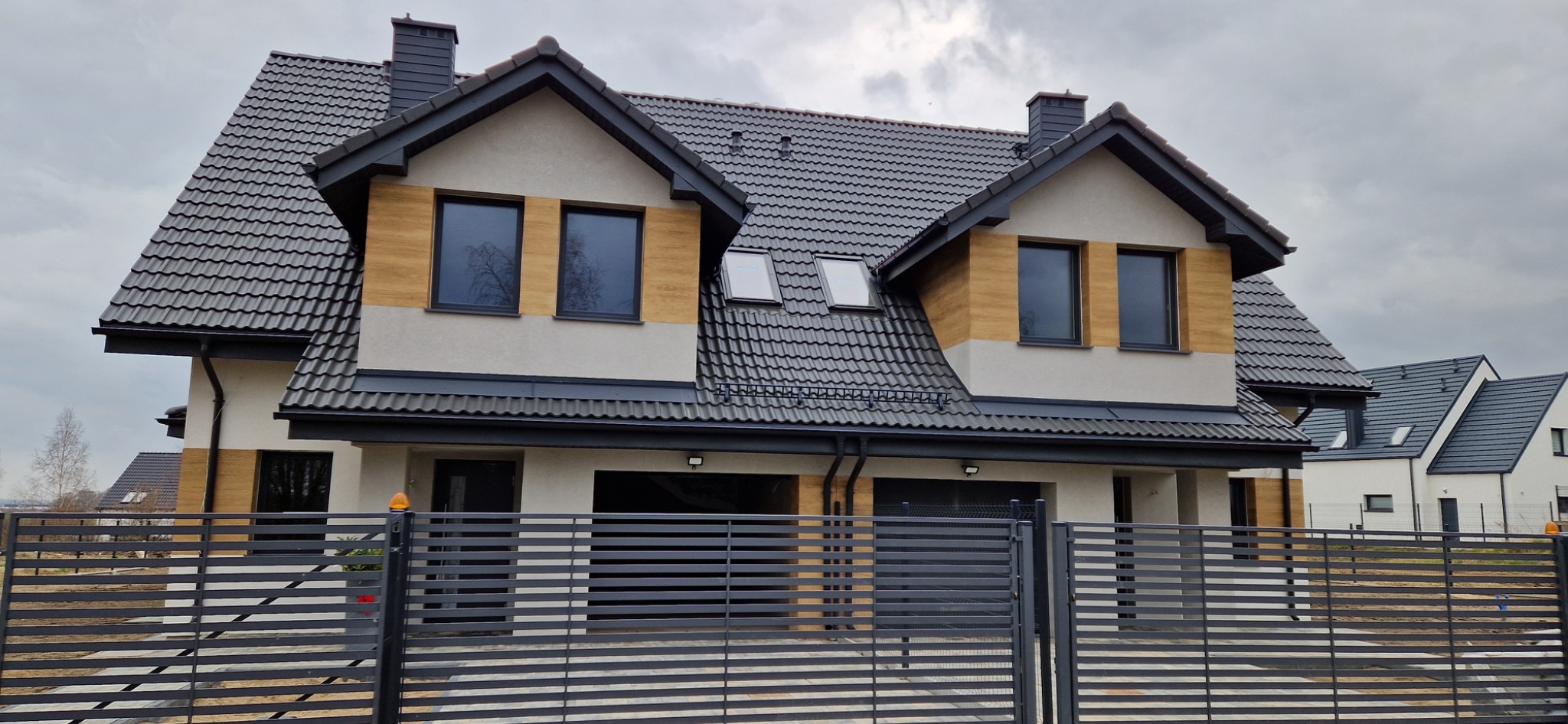 Nowoczesny dom z szarą dachówką i elementami drewnopodobnymi na elewacji, widoczny zza metalowego ogrodzenia. Uwagę zwracają okna dachowe i dwuspadowy dach.