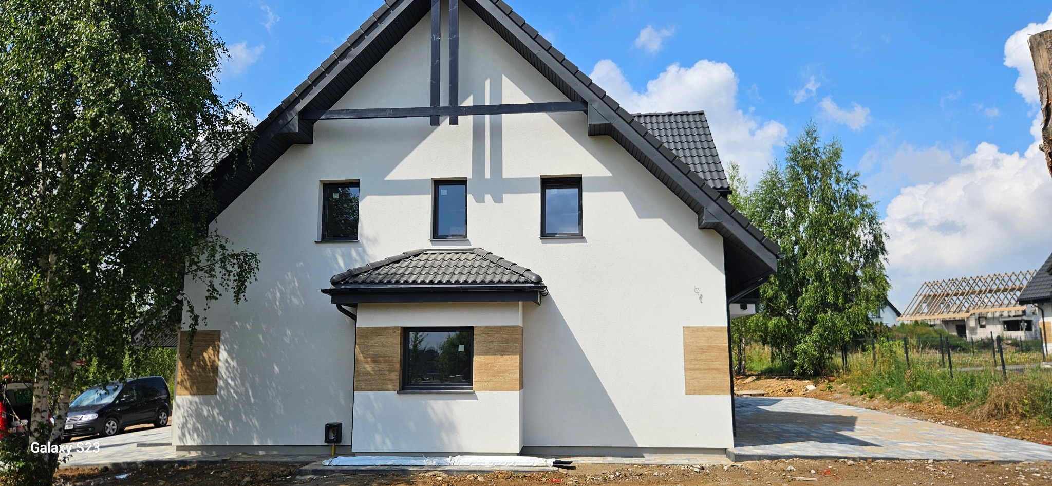 Nowoczesny dom z białą elewacją i czarnym dachem, widok z boku, z fragmentem podjazdu i zieleni. Na elewacji widoczne elementy dekoracyjne imitujące drewno.