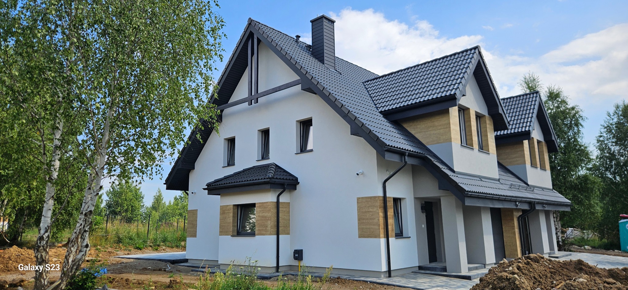 Nowoczesny dom z białą elewacją, czarnym dachem i elementami dekoracyjnymi z drewna. Widoczne okna, rynny i fragment ogrodu z drzewem. Ujęcie z boku, w słoneczny dzień.