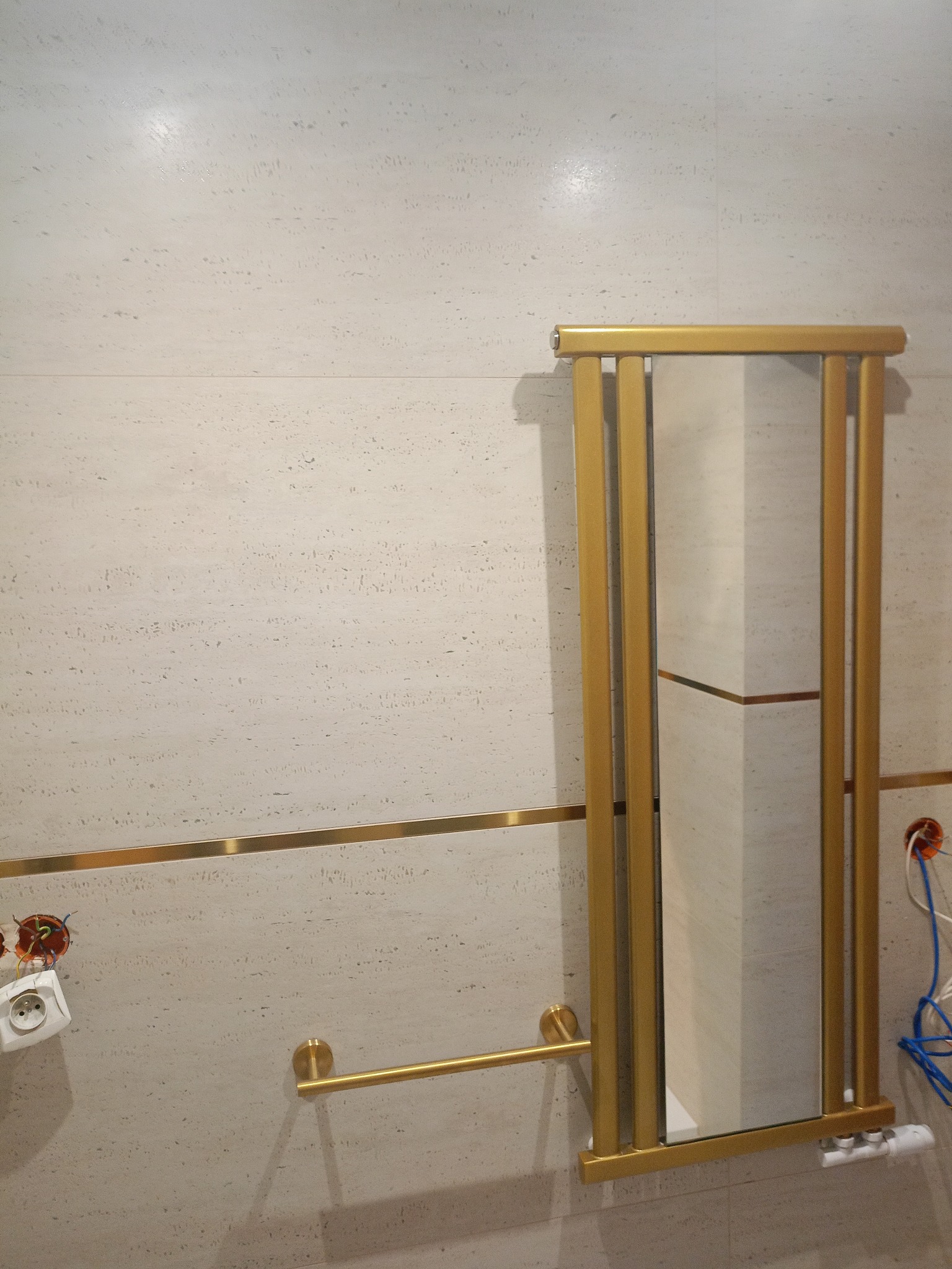 Wykończona ściana łazienki z beżowych płytek z złotymi akcentami: listwa, wieszak na ręczniki i rama lustra. Widoczne gniazdka elektryczne przed montażem.