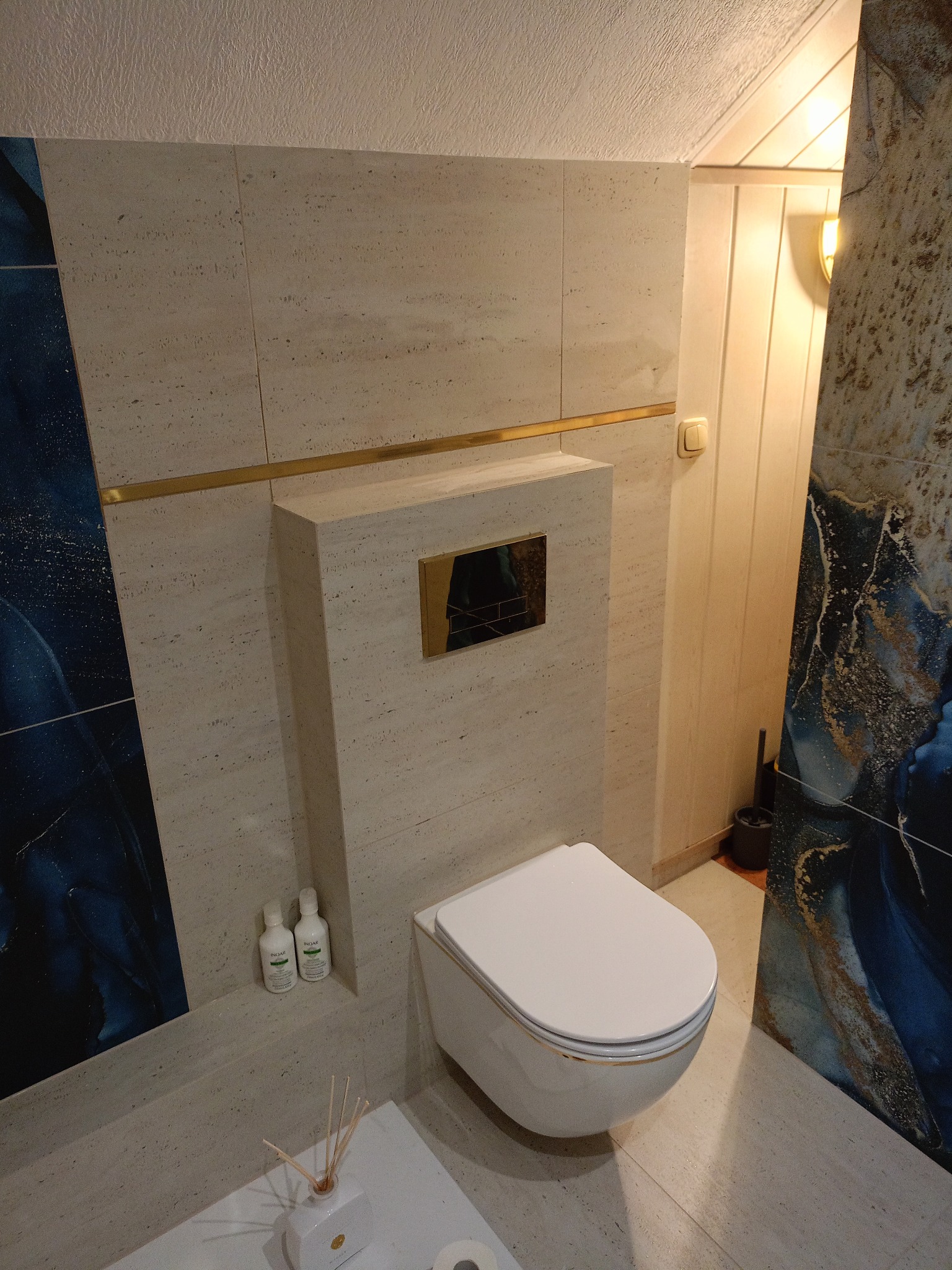 Nowoczesna toaleta podwieszana w łazience wykończonej beżowymi płytkami z delikatnym wzorem. Dekoracyjne, niebieskie panele po bokach. Złote akcenty i minimalistyczny design.