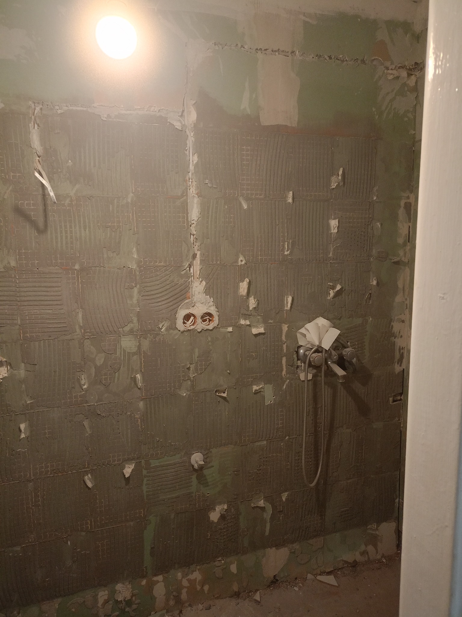 Ściana łazienki w trakcie demontażu płytek, widoczne ślady po kleju i instalacja wodna zabezpieczona papierem. Przygotowanie do dalszych prac remontowych.