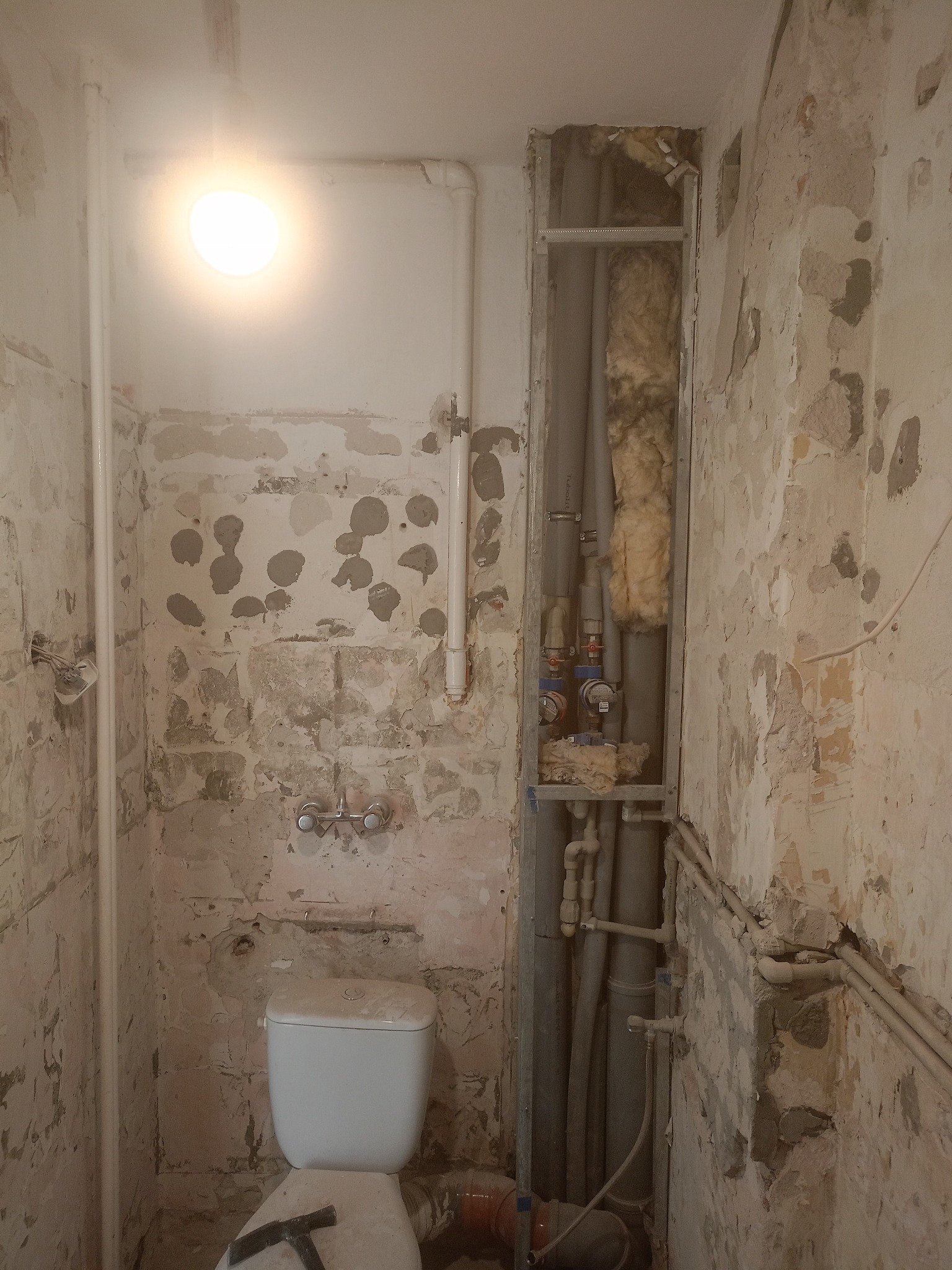 Łazienka w trakcie demontażu: odsłonięte rury w ścianie, surowe ściany z resztkami farby, wisząca toaleta i narzędzie na sedesie. Widok na wnętrze w trakcie prac.