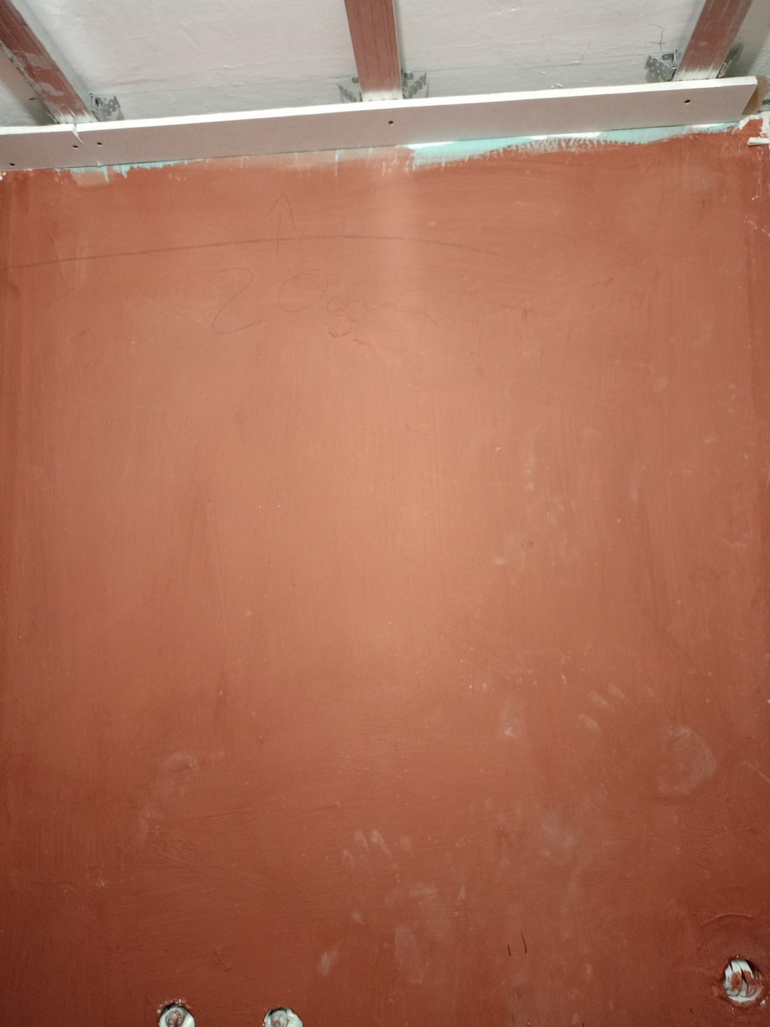 Ściana pokryta czerwoną gładzią, z widocznymi śladami po szpachlowaniu i zaznaczeniami ołówkiem. U góry widoczne elementy konstrukcji sufitu podwieszanego.