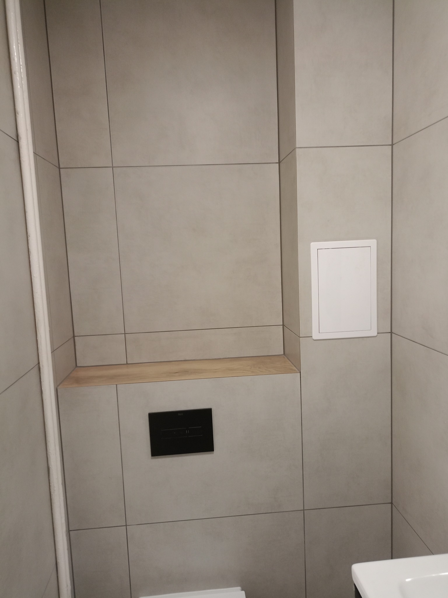 Wykończona łazienka z szarymi płytkami, drewnianą półką i czarnym przyciskiem spłuczki. Widoczny fragment umywalki. Minimalistyczny design.