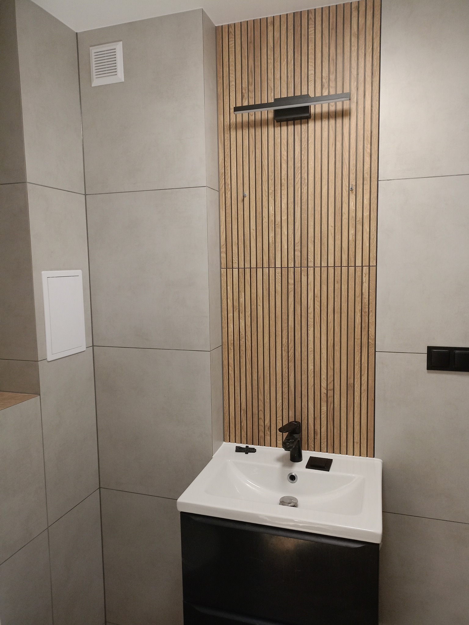 Nowoczesna łazienka z umywalką na szafce, drewnianą ścianą z lamelami i minimalistycznym oświetleniem. Szare płytki na ścianach i podłodze. Czarna bateria umywalkowa.