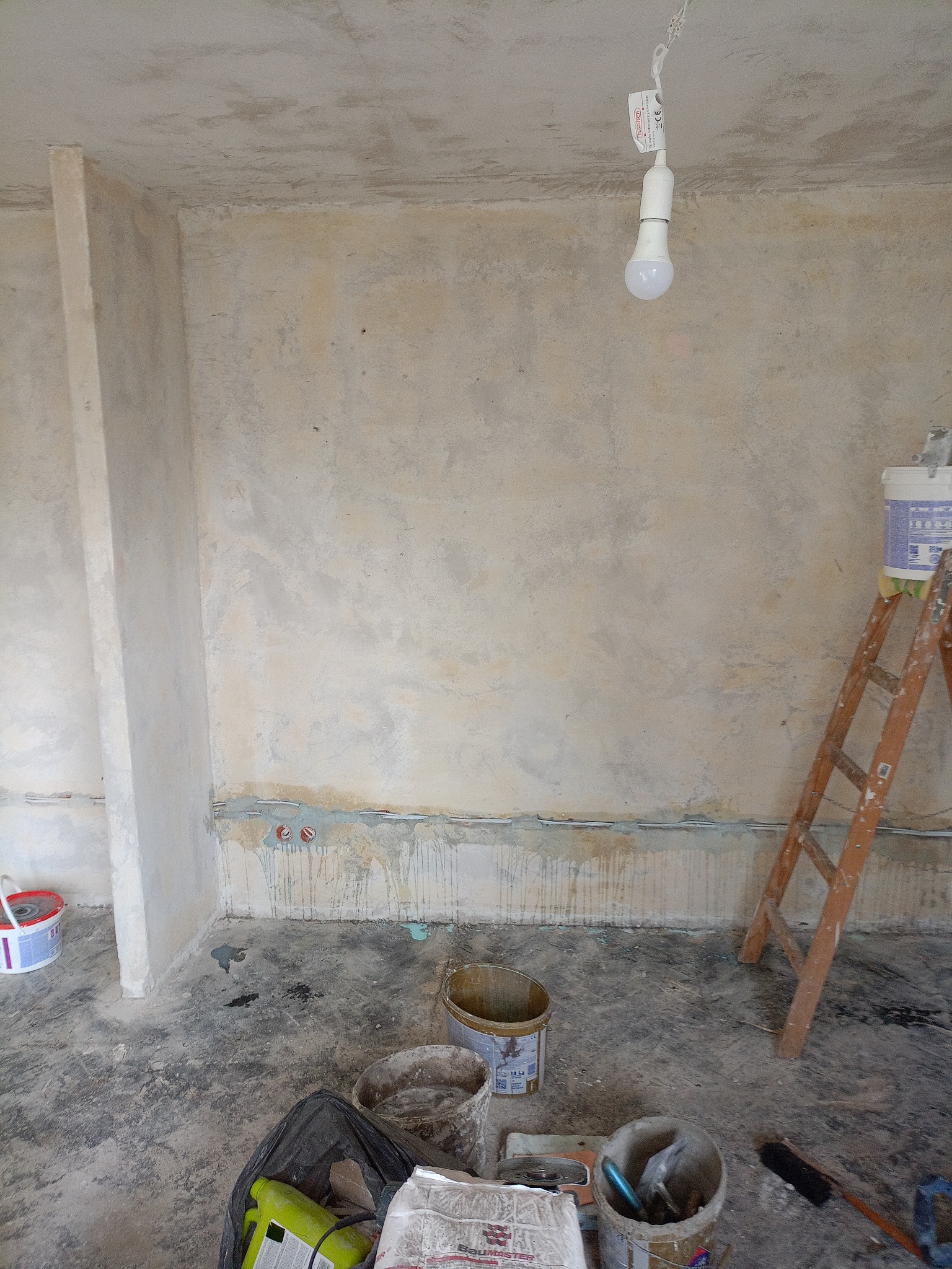 Pokój w trakcie prac wykończeniowych: surowe ściany, widoczne ślady po instalacjach, drabina oparta o ścianę, narzędzia i materiały budowlane na podłodze.