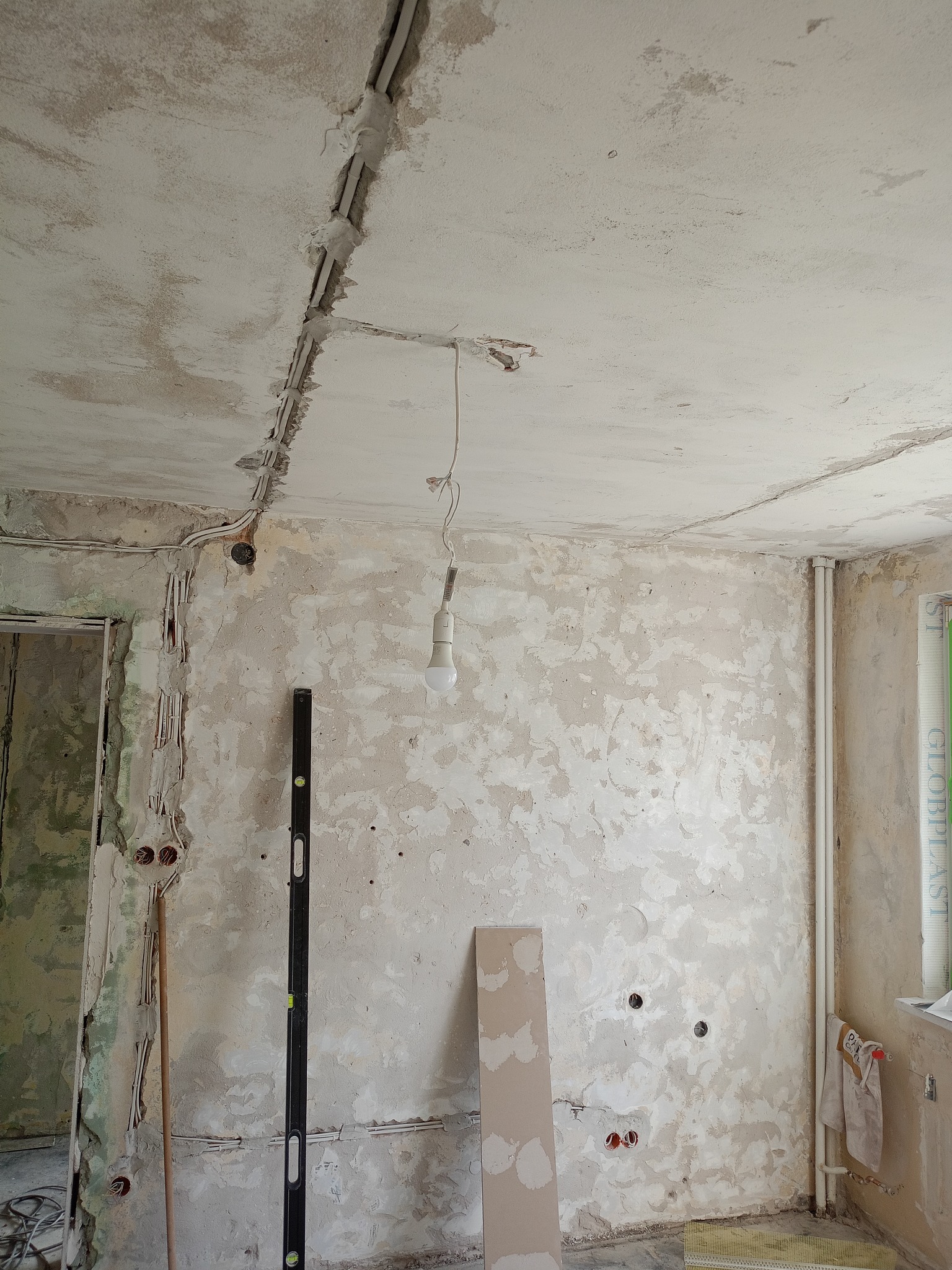 Wnętrze w trakcie remontu: odsłonięte przewody elektryczne w ścianach i suficie, wisząca żarówka, poziomica oparta o ścianę, surowe ściany przygotowane do dalszych prac.