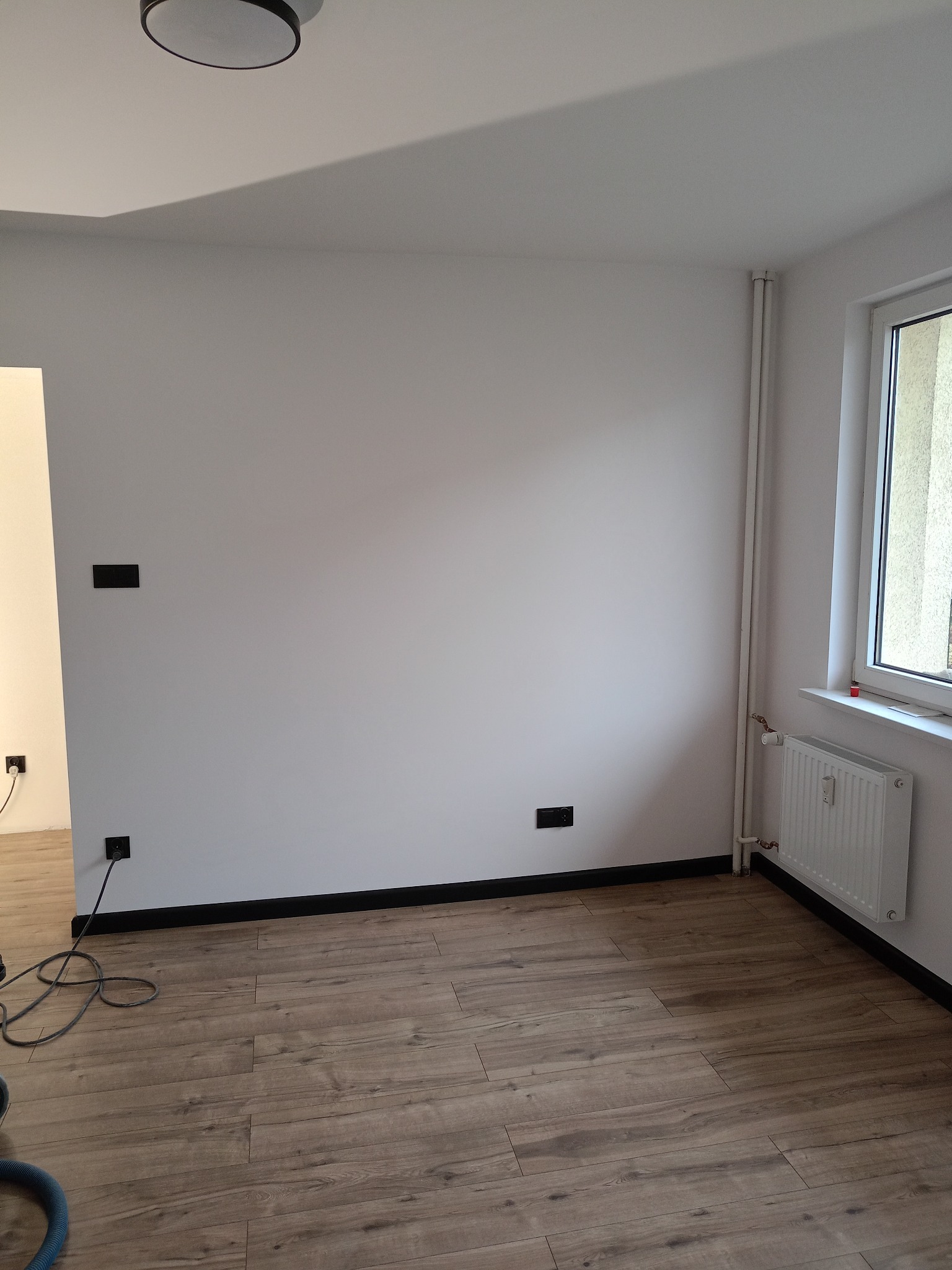 Jasne panele podłogowe w pokoju z białymi ścianami i czarnymi listwami przypodłogowymi. Grzejnik pod oknem. Widoczne gniazdka elektryczne.