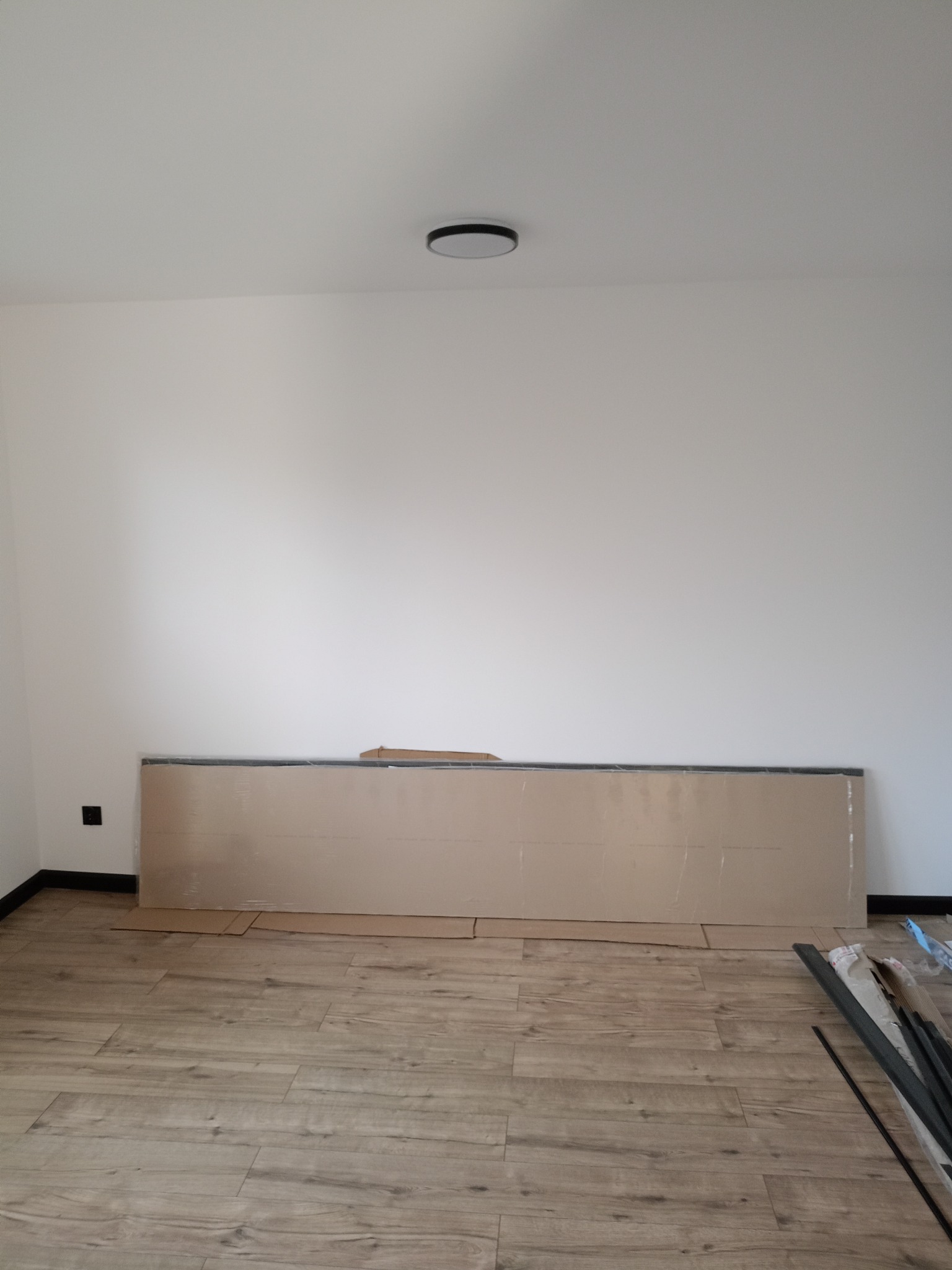 Ułożone panele podłogowe w jasnym pokoju, zabezpieczone kartonem przy ścianie. Widoczne listwy przypodłogowe i minimalistyczna lampa sufitowa.