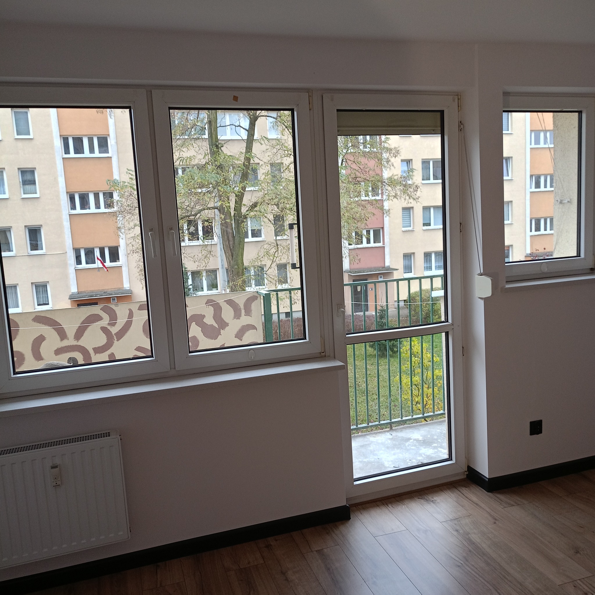 Jasne wnętrze po remoncie: panele podłogowe, białe ściany, okna z widokiem na blok i balkon. Nowoczesny grzejnik pod oknem. Wykończenie wnętrz w neutralnej kolorystyce.