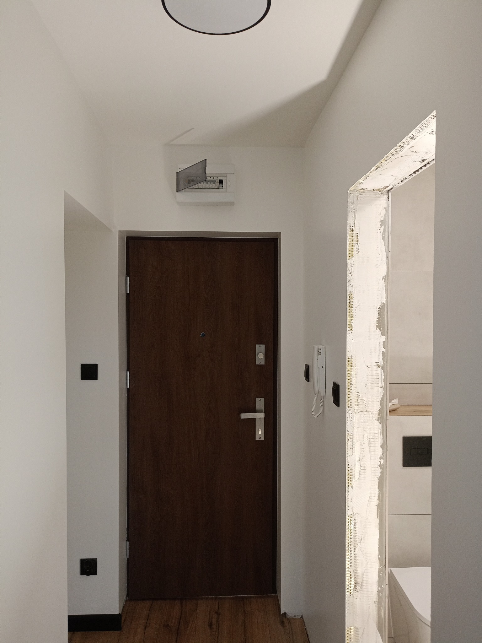 Wnętrze w trakcie prac: widok na brązowe drzwi wejściowe, białe ściany z osprzętem elektrycznym i fragment łazienki z widoczną toaletą przez ościeżnicę w trakcie wykańczania.