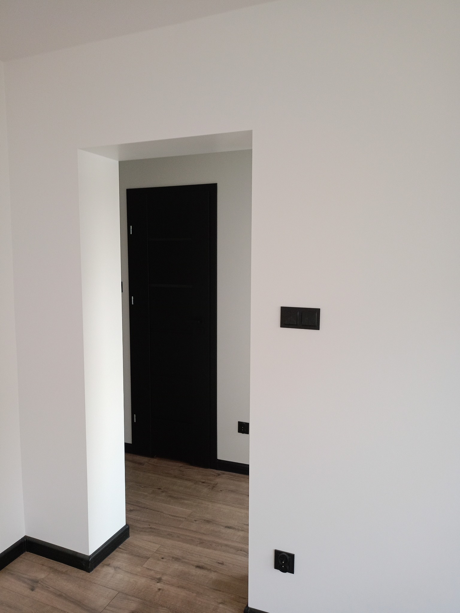 Białe ściany z czarnymi akcentami: listwy przypodłogowe, drzwi i gniazdka. Podłoga z paneli imitujących drewno. Minimalistyczne wykończenie wnętrza.
