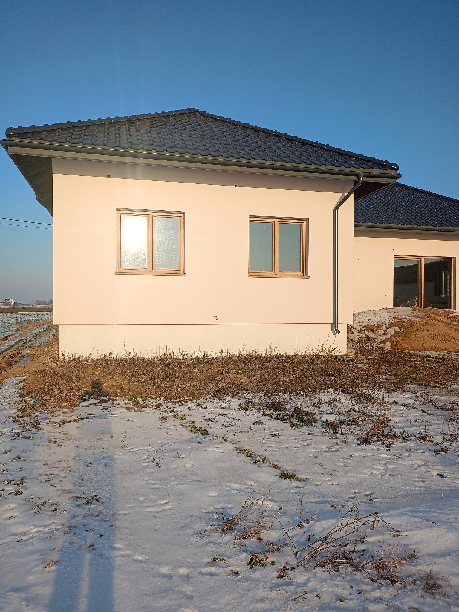 Jasny, otynkowany dom jednorodzinny z ciemnym dachem w zimowej scenerii. Dwa okna z drewnianymi ramami odbijają światło słoneczne. Częściowo ośnieżone pole w tle.