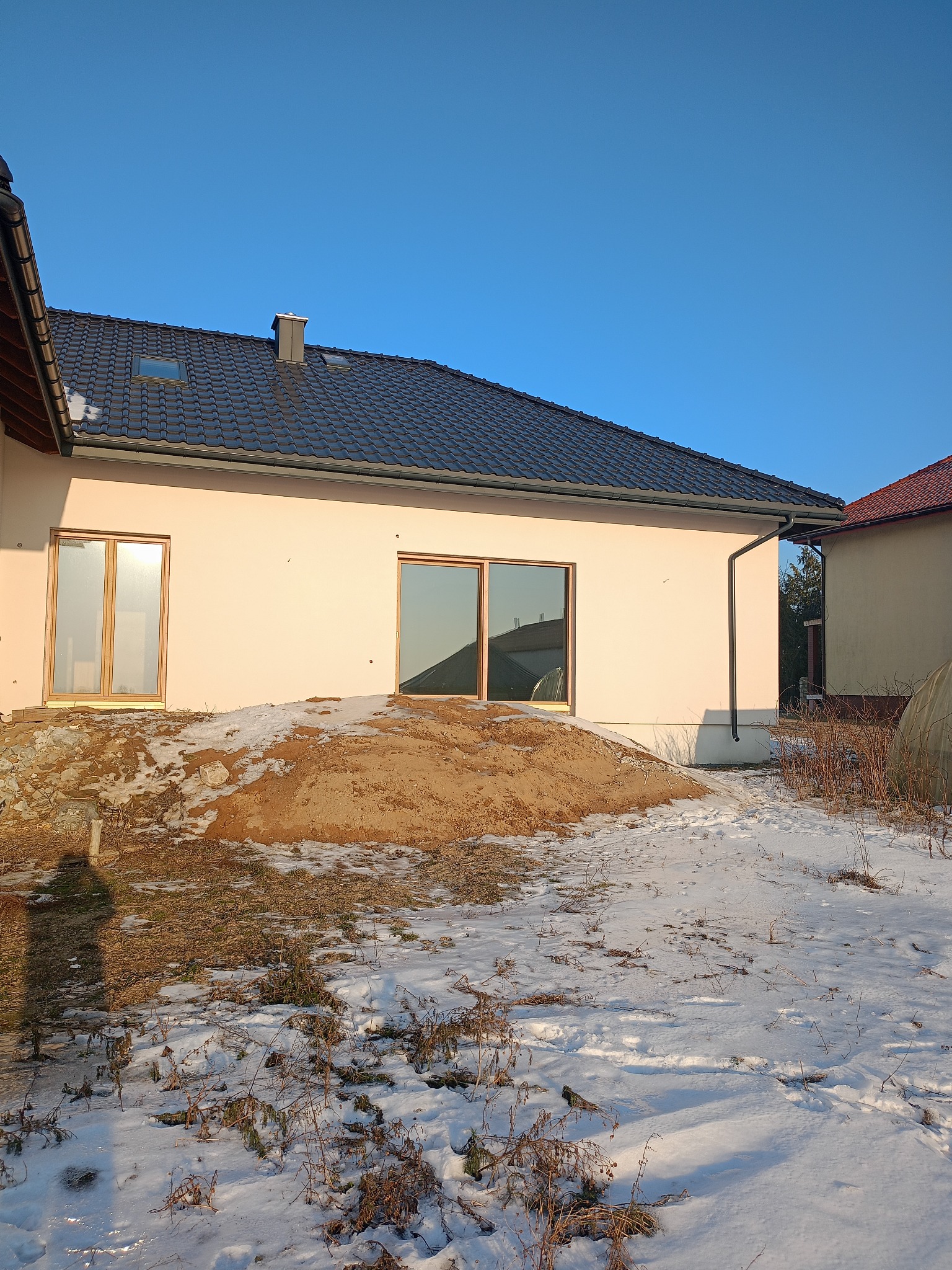 Jasny, parterowy dom z ciemną dachówką w zimowej scenerii. Świeżo otynkowana elewacja, widoczne okna z drewnianą ramą i zasypany śniegiem teren.