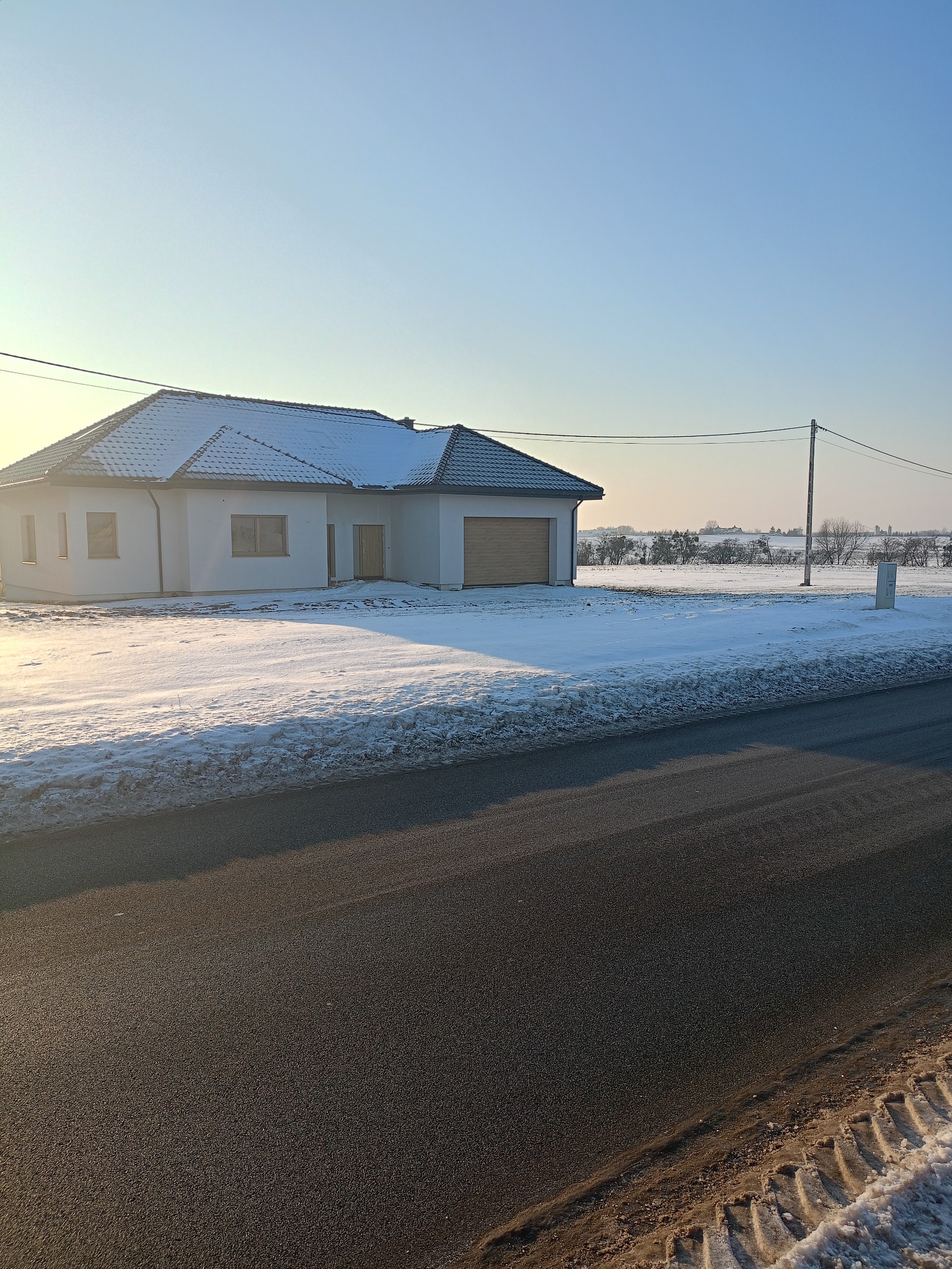 Parterowy dom z garażem w zimowej scenerii, pokryty śniegiem dach i otoczenie. Widok z poziomu ulicy, w tle pole i drzewa. Jasne, zimowe światło.