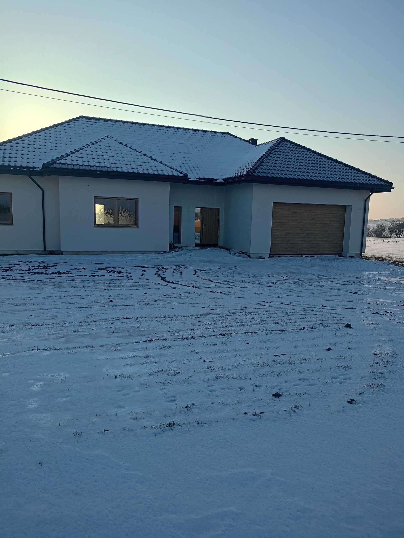 Nowoczesny, parterowy dom z garażem w zimowej scenerii. Biała elewacja kontrastuje z ciemną dachówką. Ślady na śniegu prowadzą do wejścia.