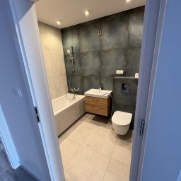 Skfuturehome - Nowoczesna łazienka z wanną, umywalką z drewnianą szafką i toaletą. Ciemne płytki na ścianie kontrastują z jasną podłogą. Widok zza otwartych drzwi.