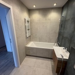 Skfuturehome - Nowoczesna łazienka w szarościach z wanną, umywalką z szafką i grzejnikiem drabinkowym. Ściany i podłoga wyłożone płytkami w odcieniach szarości. Widoczne oświetlenie punktowe.