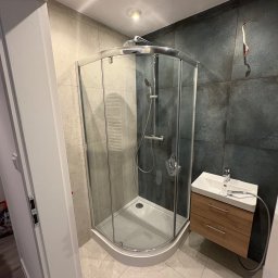 Skfuturehome - Nowoczesna kabina prysznicowa narożna z umywalką na szafce w wykończonej łazience. Widoczne płytki w odcieniach szarości i beżu, oświetlenie punktowe.