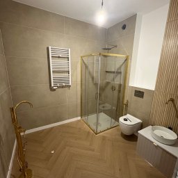 Skfuturehome - Nowoczesna łazienka z kabiną prysznicową w złotych ramach, grzejnikiem drabinkowym i umywalką nablatową. Parkiet ułożony w jodełkę. Widoczne elementy złotej armatury.