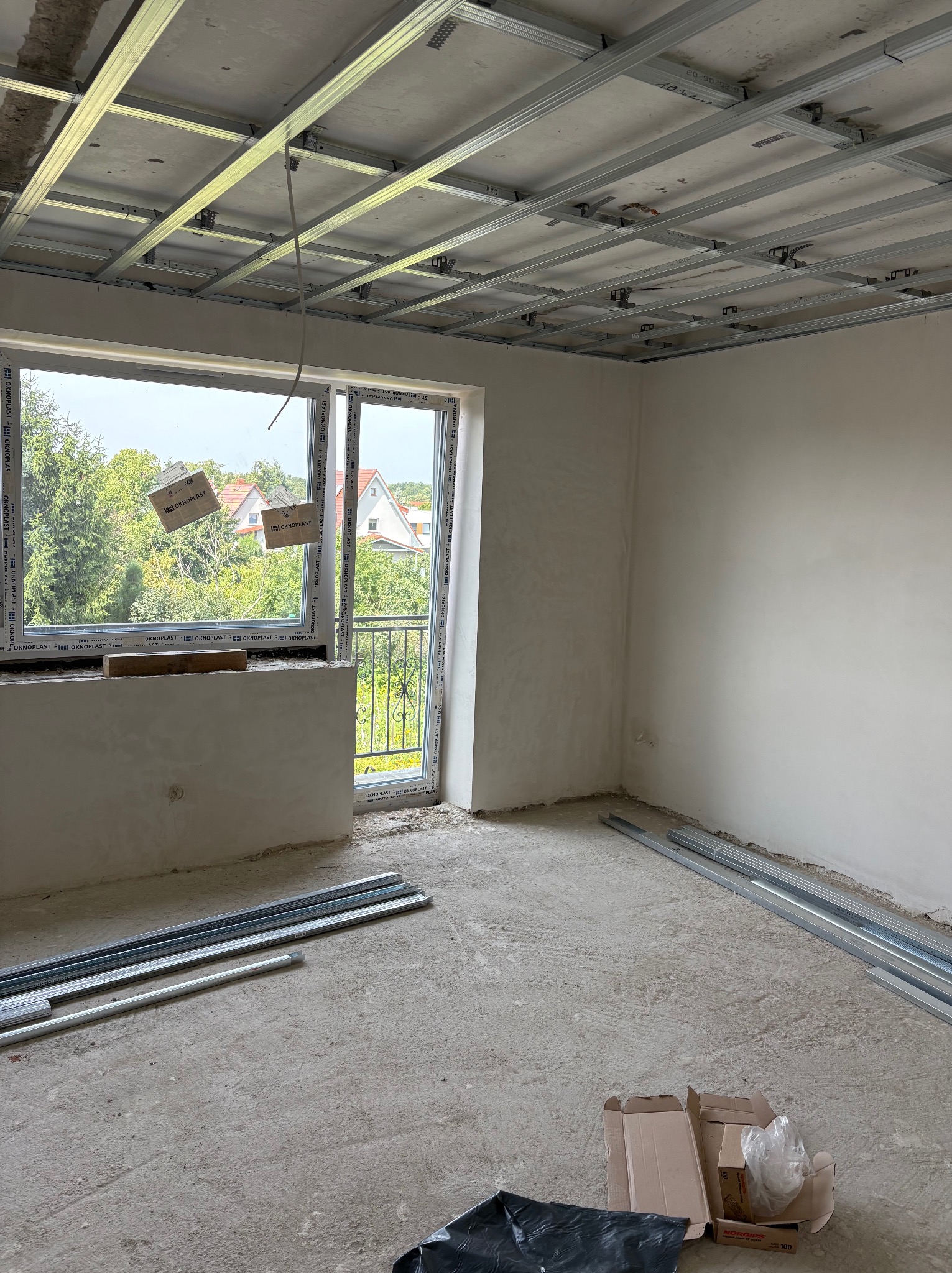 Wnętrze w trakcie prac: metalowa konstrukcja sufitu podwieszanego, surowe ściany, okno z widokiem na zieleń. Na podłodze profile i karton. Etap przed wykończeniem.