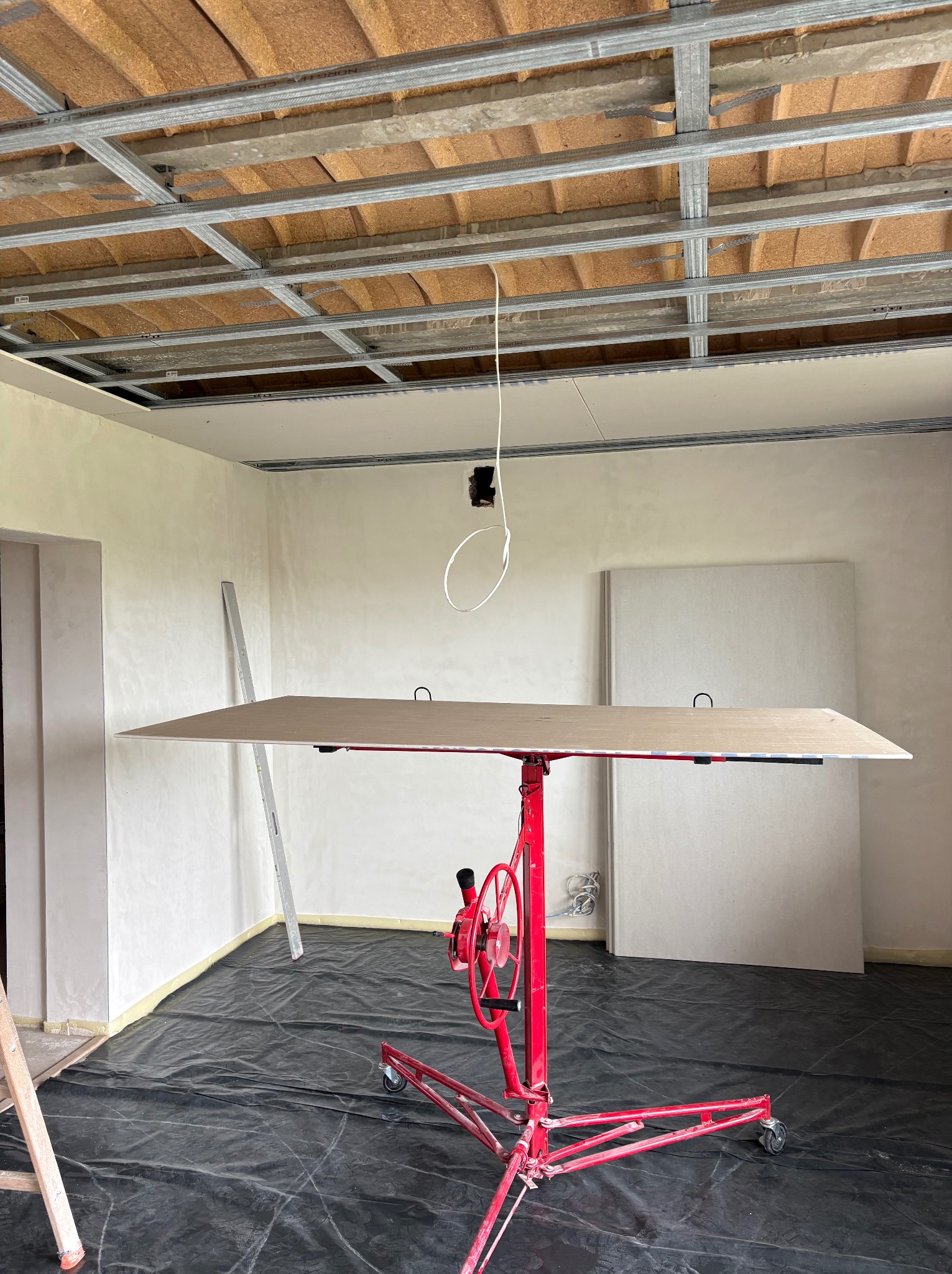 Montaż płyty gipsowej na suficie za pomocą podnośnika. Widoczne metalowe profile sufitowe i przygotowane płyty. Podłoga zabezpieczona folią.