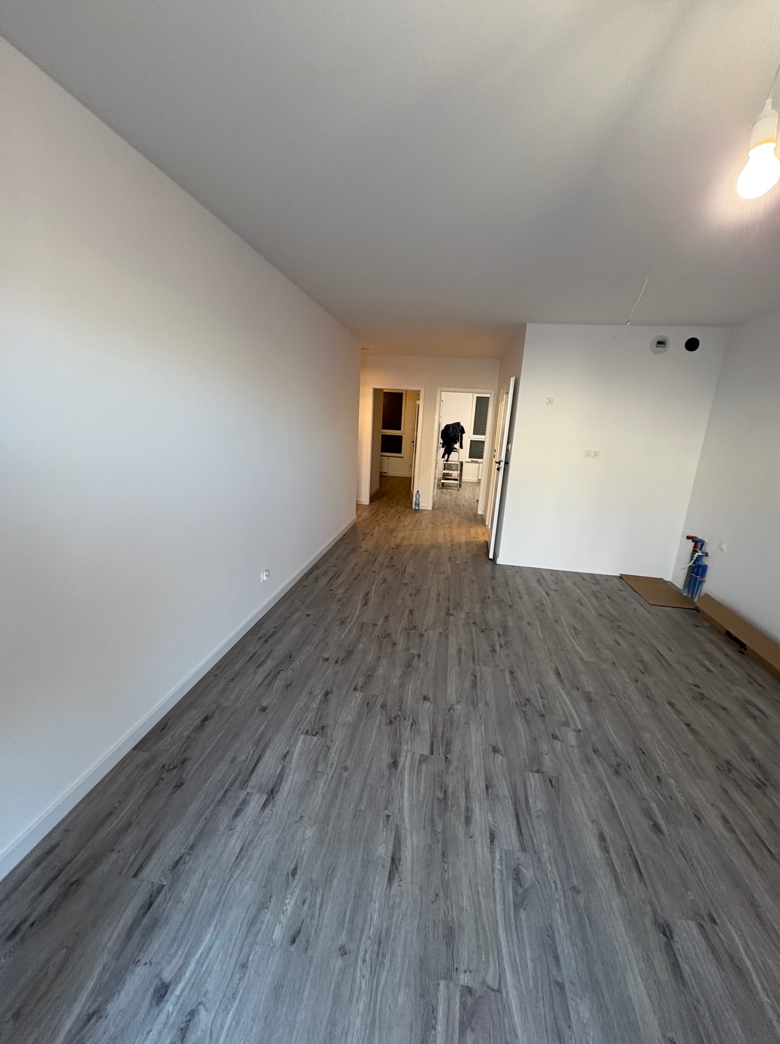 Puste, jasne wnętrze po remoncie z szarymi panelami podłogowymi. Widoczne białe ściany, sufit z lampą i otwarte drzwi do kolejnych pomieszczeń.