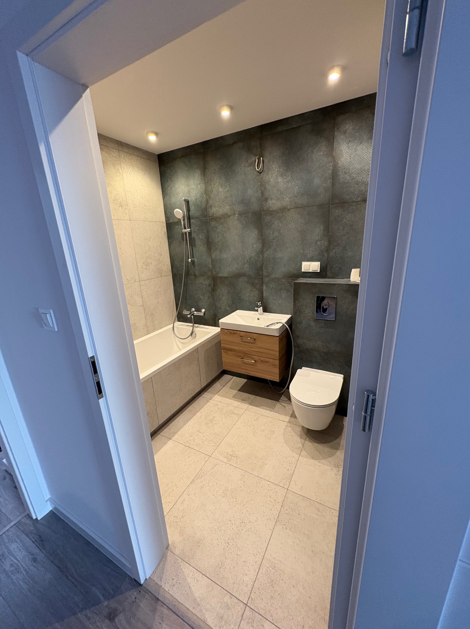 Nowoczesna łazienka z wanną, umywalką z drewnianą szafką i toaletą. Ciemne płytki na ścianie kontrastują z jasną podłogą. Widok zza otwartych drzwi.