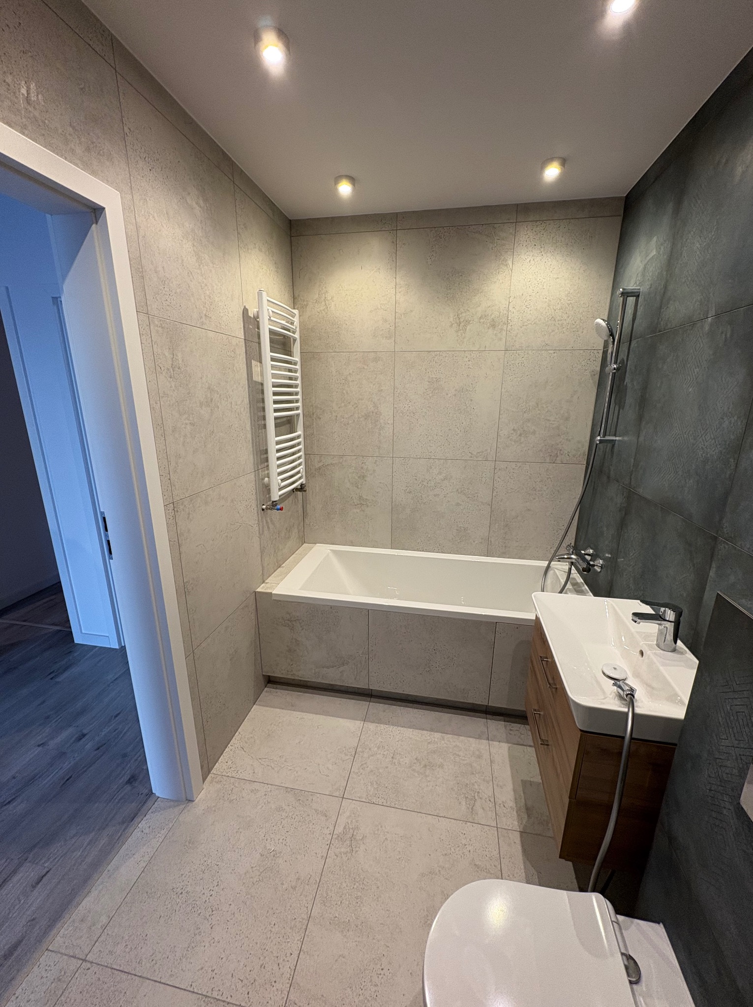 Nowoczesna łazienka w szarościach z wanną, umywalką z szafką i grzejnikiem drabinkowym. Ściany i podłoga wyłożone płytkami w odcieniach szarości. Widoczne oświetlenie punktowe.