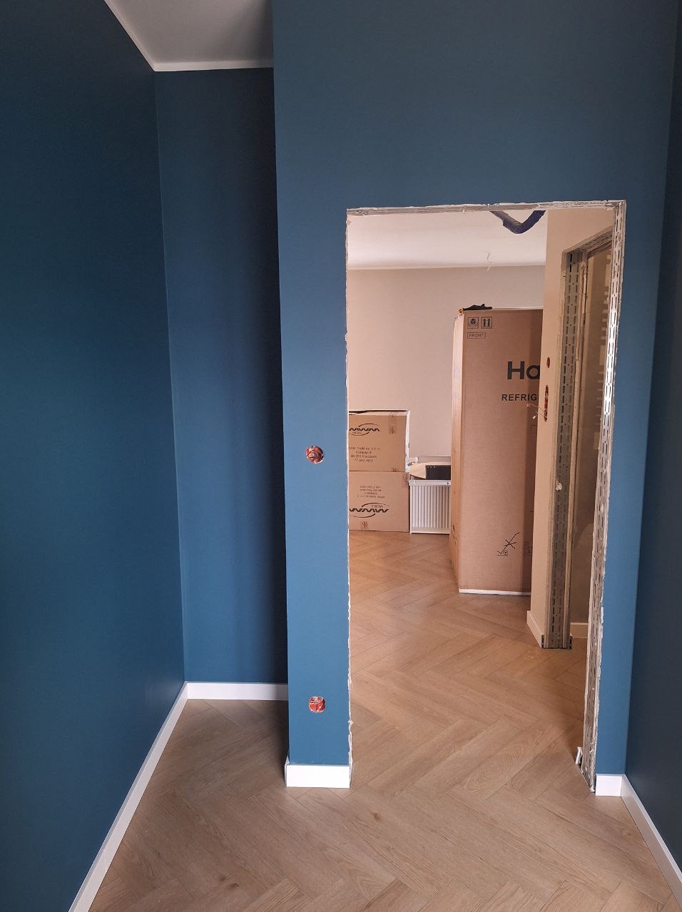 Ściana pomalowana na granatowo, parkiet w jodełkę, widok przez otwór drzwiowy na kartony i grzejnik. Gładkie ściany, listwy przypodłogowe.