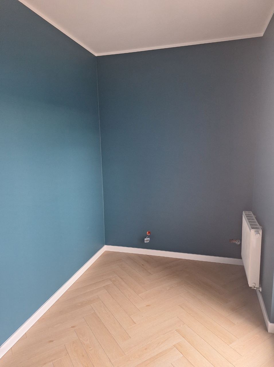 Pomalowane na dwa odcienie niebieskiego ściany w rogu pokoju z parkietem ułożonym w jodełkę. Białe listwy przypodłogowe i grzejnik.
