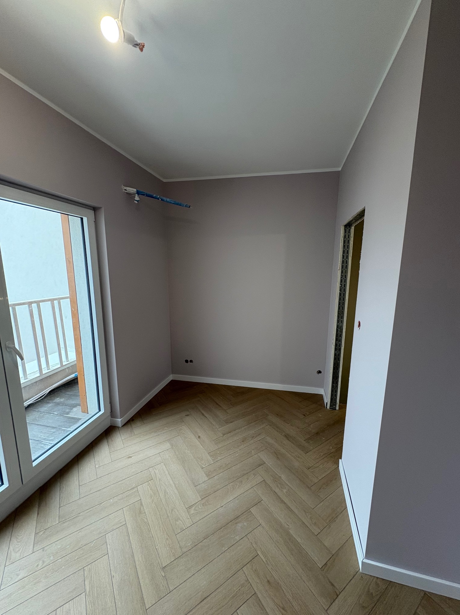 Pokój po malowaniu ścian na liliowo-szaro z parkietem w jodełkę. Widoczne gniazdka, listwy przypodłogowe i fragment balkonu przez otwarte drzwi balkonowe.