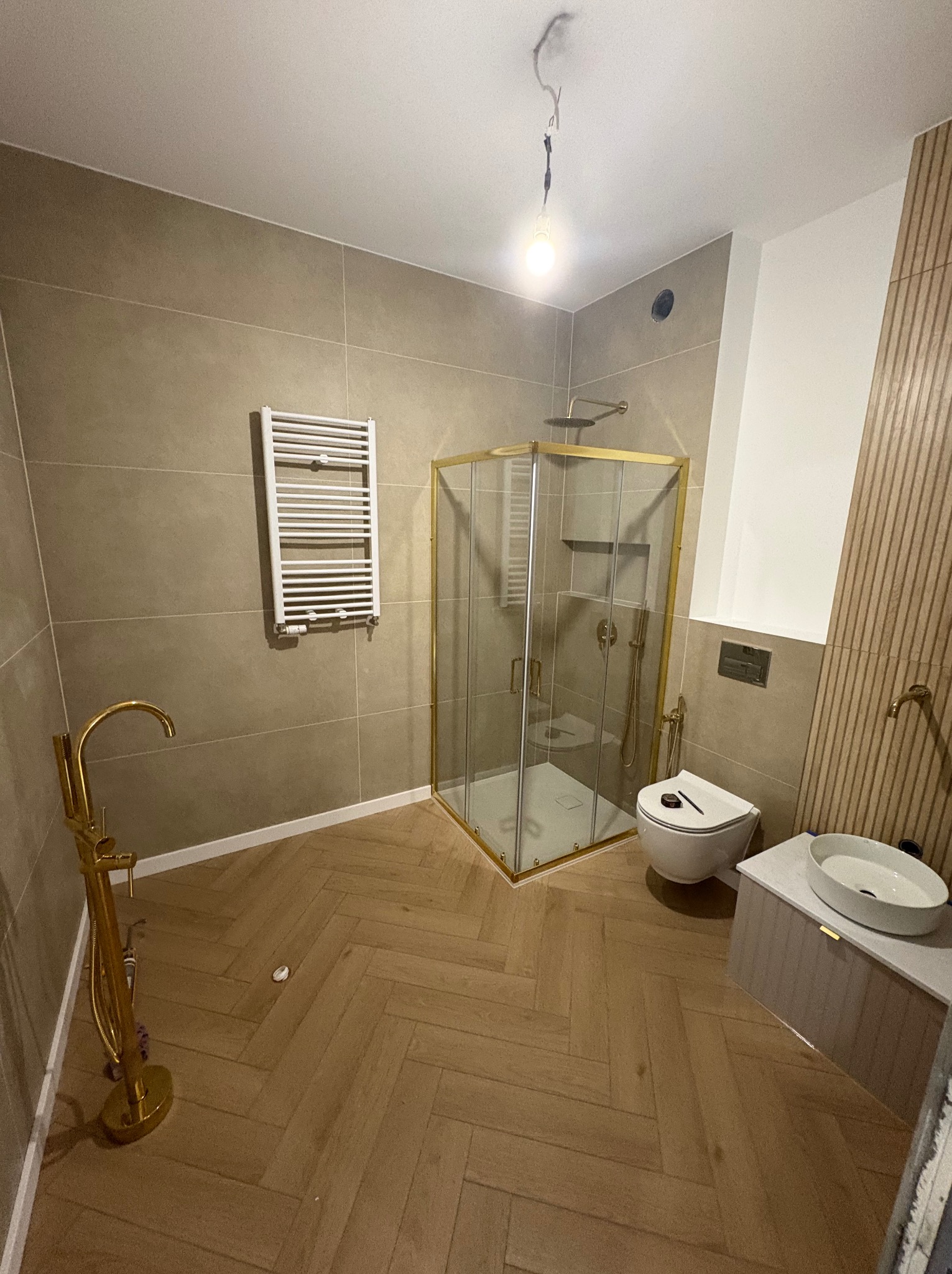 Nowoczesna łazienka z kabiną prysznicową w złotych ramach, grzejnikiem drabinkowym i umywalką nablatową. Parkiet ułożony w jodełkę. Widoczne elementy złotej armatury.