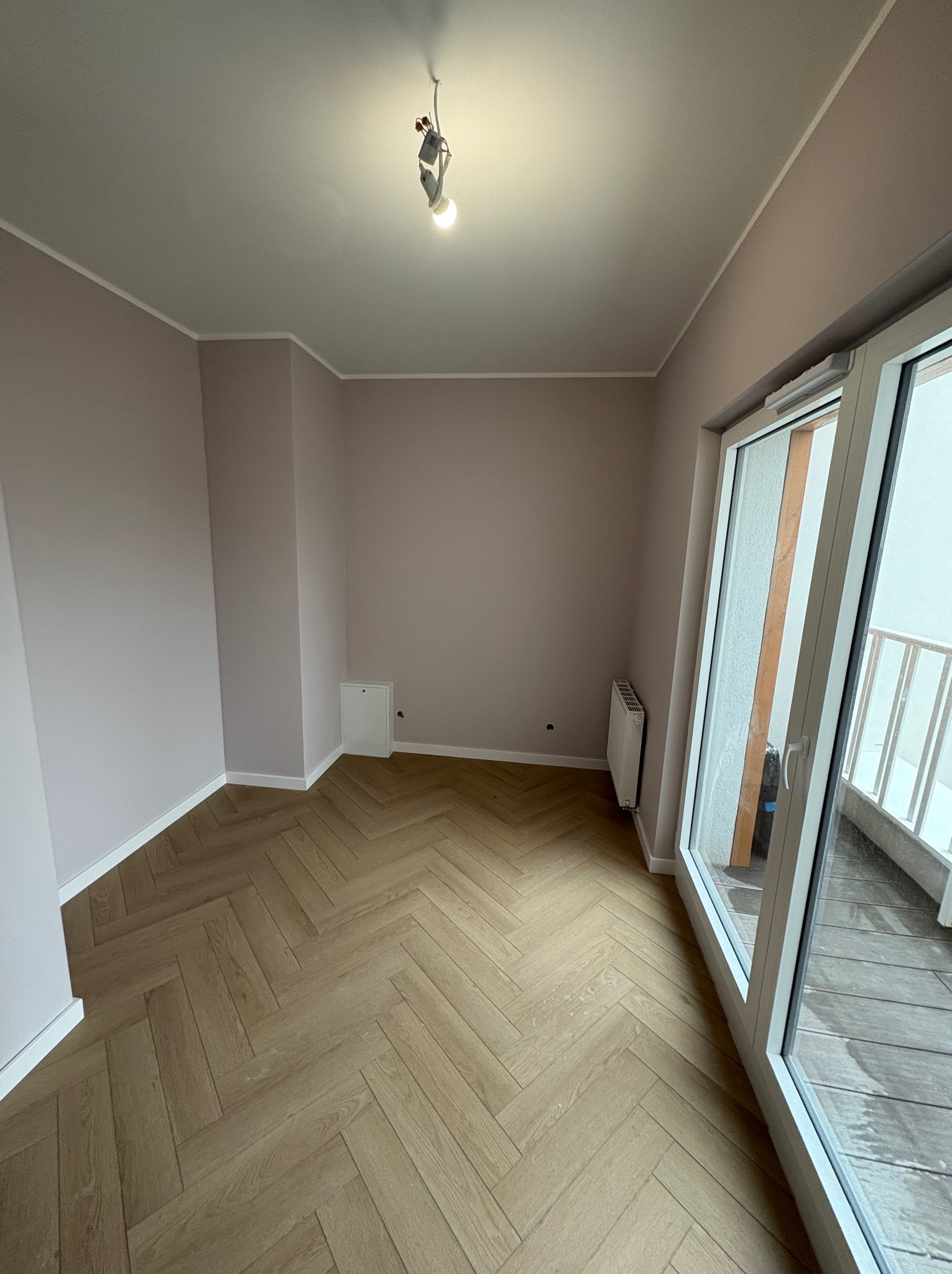 Jasne, odnowione wnętrze z parkietem w jodełkę, świeżo pomalowanymi ścianami w odcieniach beżu i dużym oknem balkonowym. Widoczny grzejnik i niedokończone oświetlenie sufitowe.