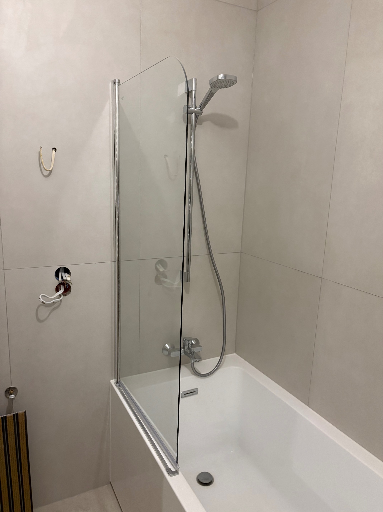 Biała wanna z zamontowaną szklaną ścianką prysznicową i chromowaną armaturą na tle jasnych, kwadratowych płytek. Widoczne niedokończone podłączenia.