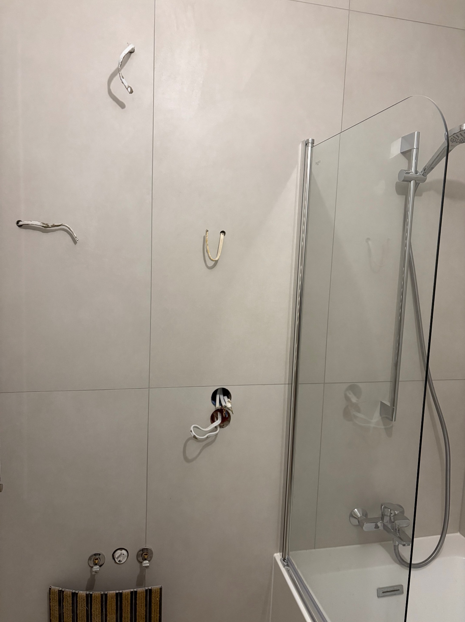 Ściana w łazience z szarymi płytkami, widoczne otwory po armaturze, wystające kable i fragment kabiny prysznicowej. Etap prac podczas wykańczania łazienki.