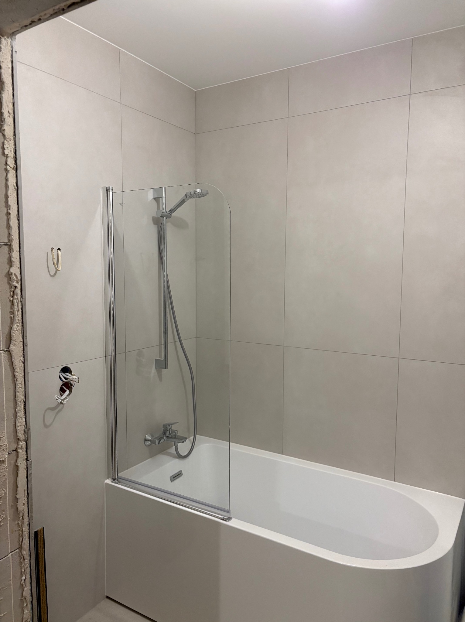 Wykończona łazienka z wanną i szklanym parawanem. Widoczne płytki w odcieniach szarości i fragment ściany w trakcie wykończenia. Minimalistyczny design.