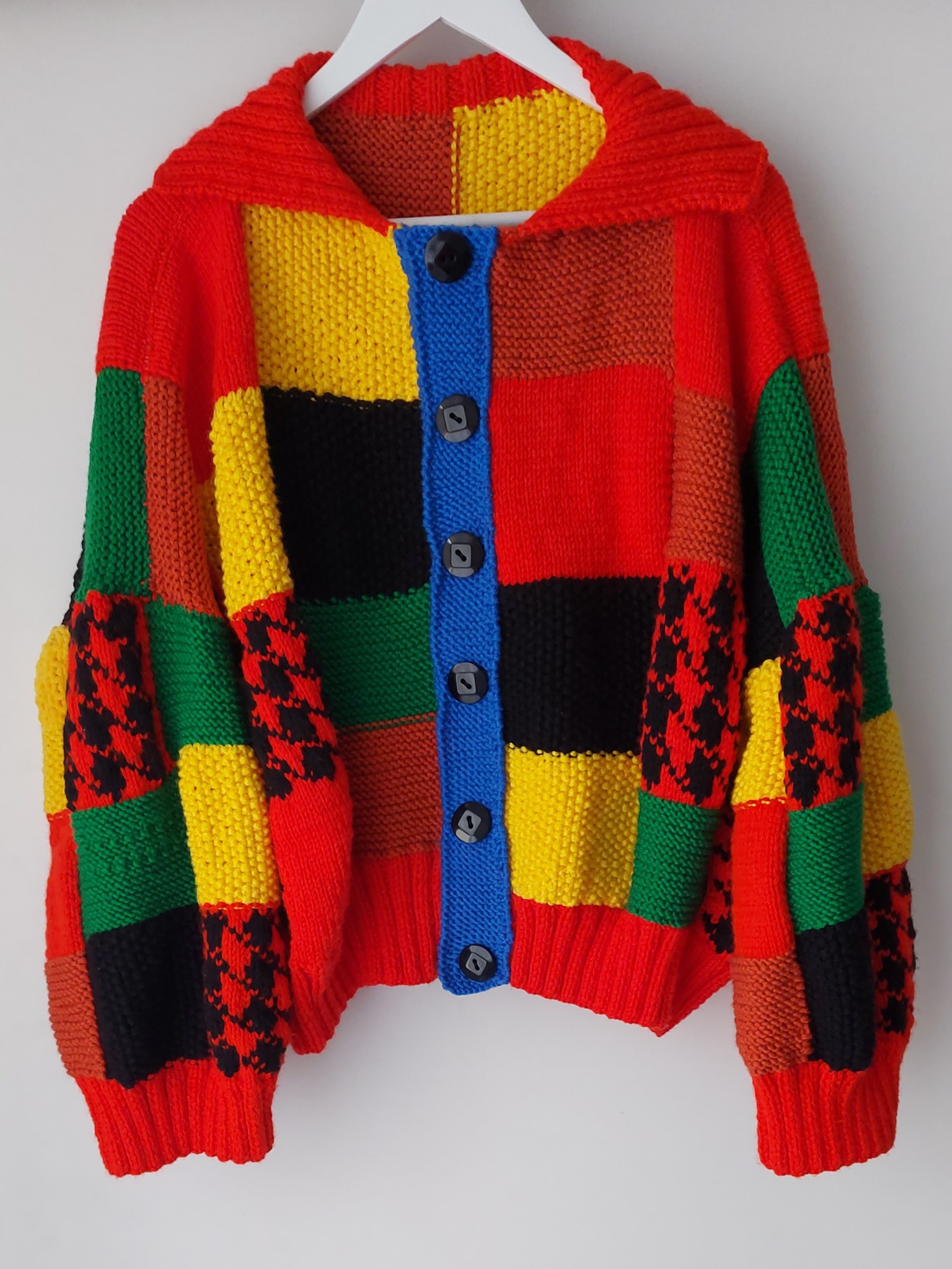 Ręcznie robiony, wielokolorowy kardigan z grubych włóczek, wiszący na białym wieszaku na tle białej ściany, prezentujący patchworkowy wzór z elementami w kolorach czerwonym, żółtym, zielonym...