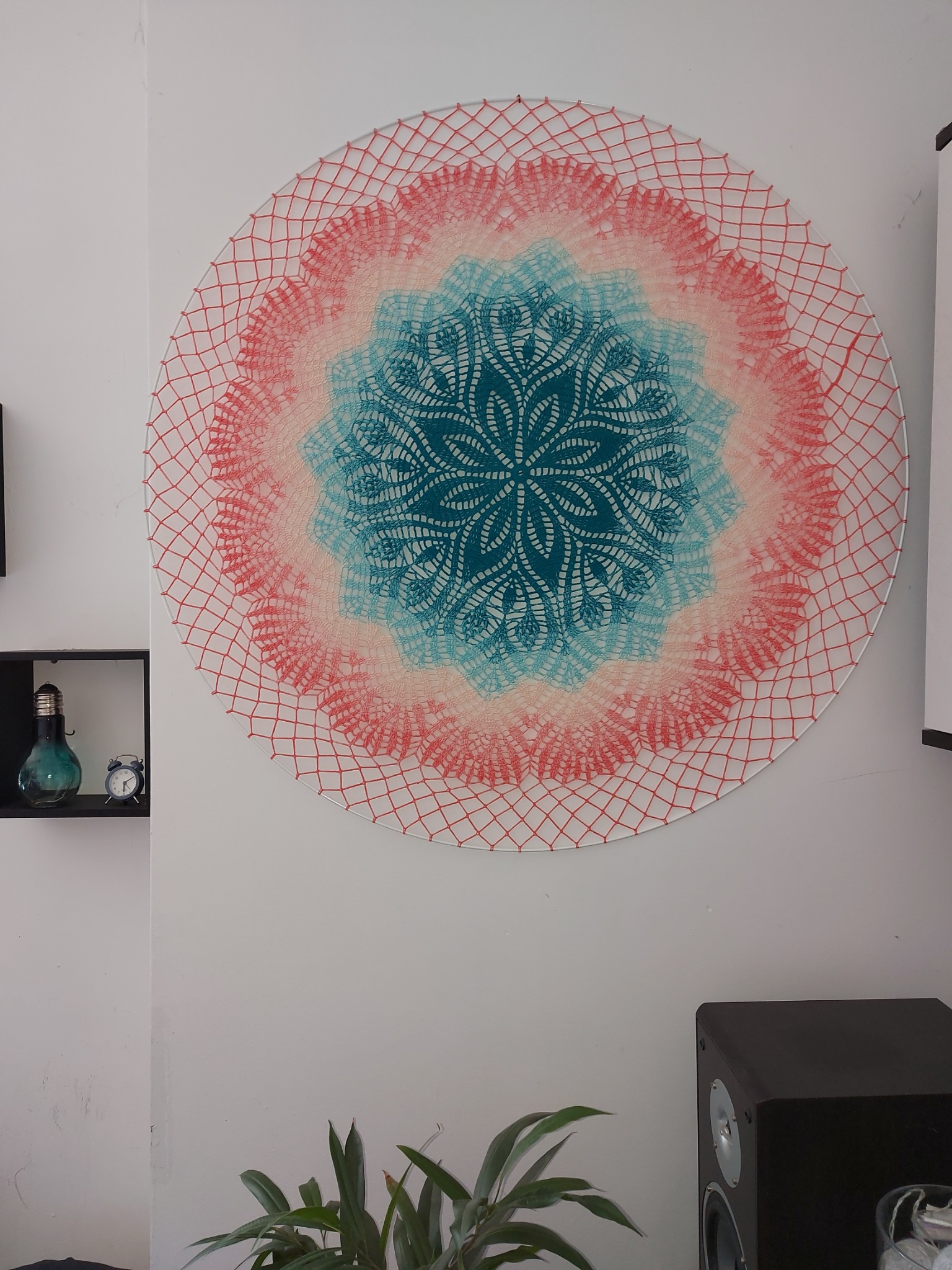 Ręcznie robiona, okrągła dekoracja ścienna z nici, w odcieniach czerwieni, różu i turkusu, zawieszona na białej ścianie, obok rośliny doniczkowej i głośnika.