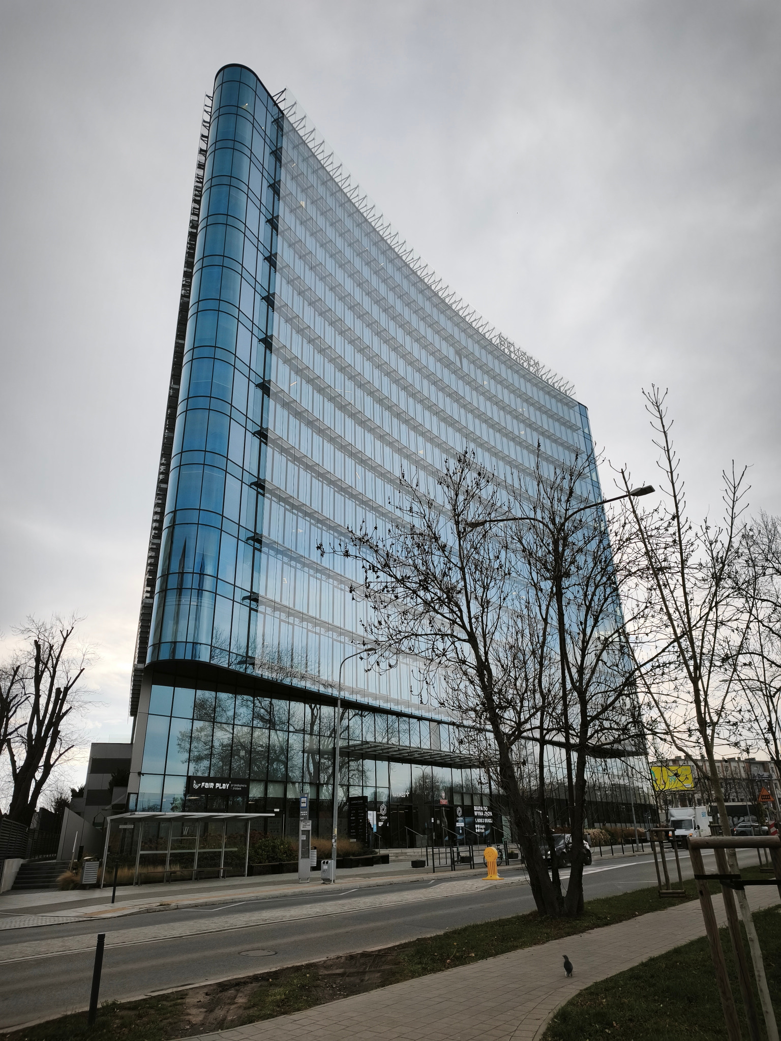 Nowoczesny, przeszklony biurowiec o zaokrąglonych narożnikach, widziany z dołu, z elementami miejskiej infrastruktury i drzewami na pierwszym planie.