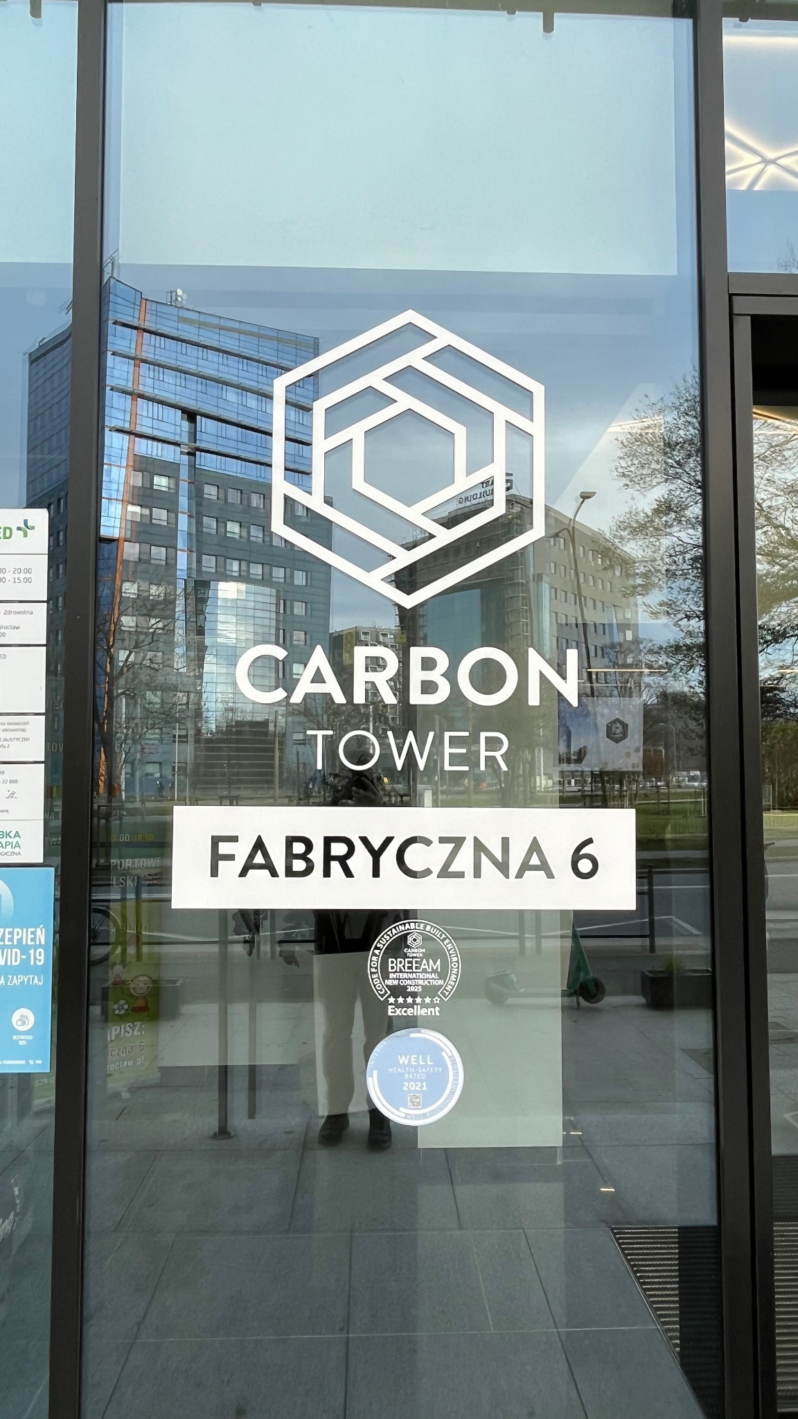 Wejście do Carbon Tower przy Fabrycznej 6, z widocznym logo i certyfikatami BREEAM oraz WELL. W tle odbicie nowoczesnego budynku.