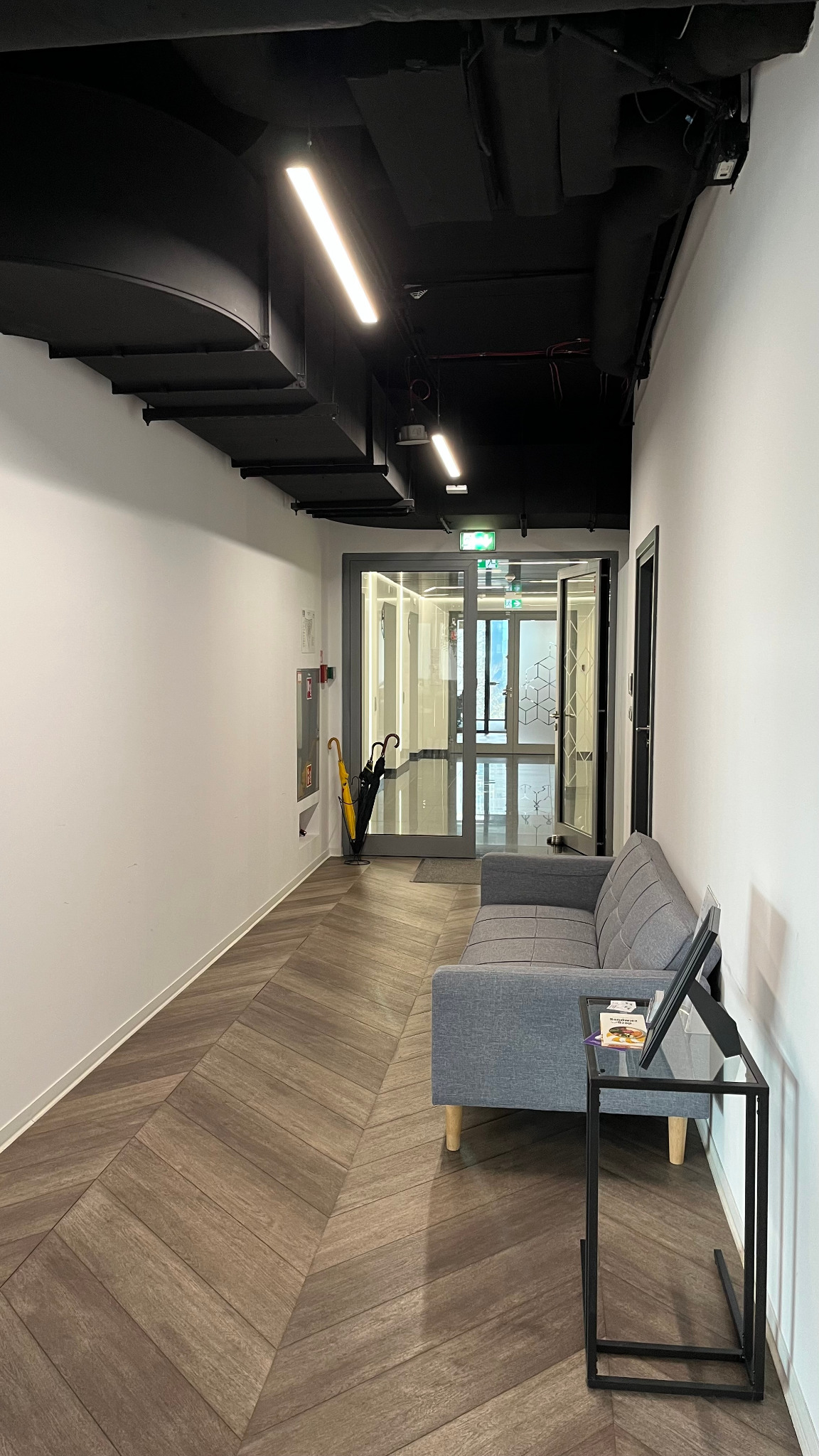 Nowoczesny hol z szarą sofą i stolikiem, parkiet w jodełkę, czarne elementy sufitu i oświetlenie liniowe. Widoczne parasole i przeszklone drzwi.