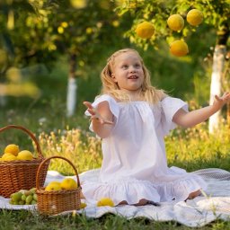 Elvira Dovhopola - Uśmiechnięta blond dziewczynka w białej sukience żongluje cytrynami na pikniku w sadzie. Kosze z owocami, miękkie światło, radosna atmosfera.