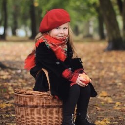 Elvira Dovhopola - Uśmiechnięta dziewczynka w czerwonym berecie i szaliku siedzi na wiklinowym koszu w jesiennym parku, trzymając jabłko. Ubrana w czarny płaszczyk i czarne buty.