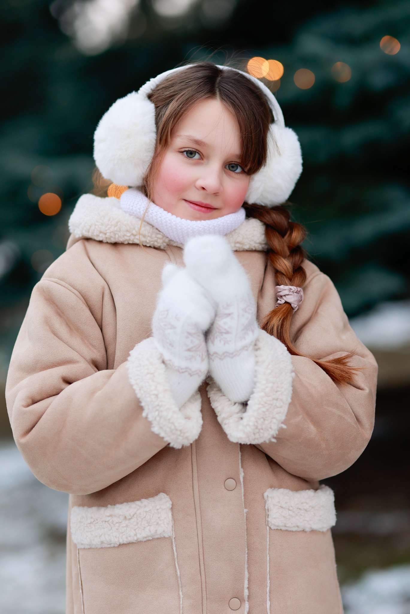 Uśmiechnięta dziewczynka w zimowej stylizacji: futrzane nauszniki, płaszcz z kożuszkiem i rękawiczki w śnieżynki, na tle choinki z bokeh.