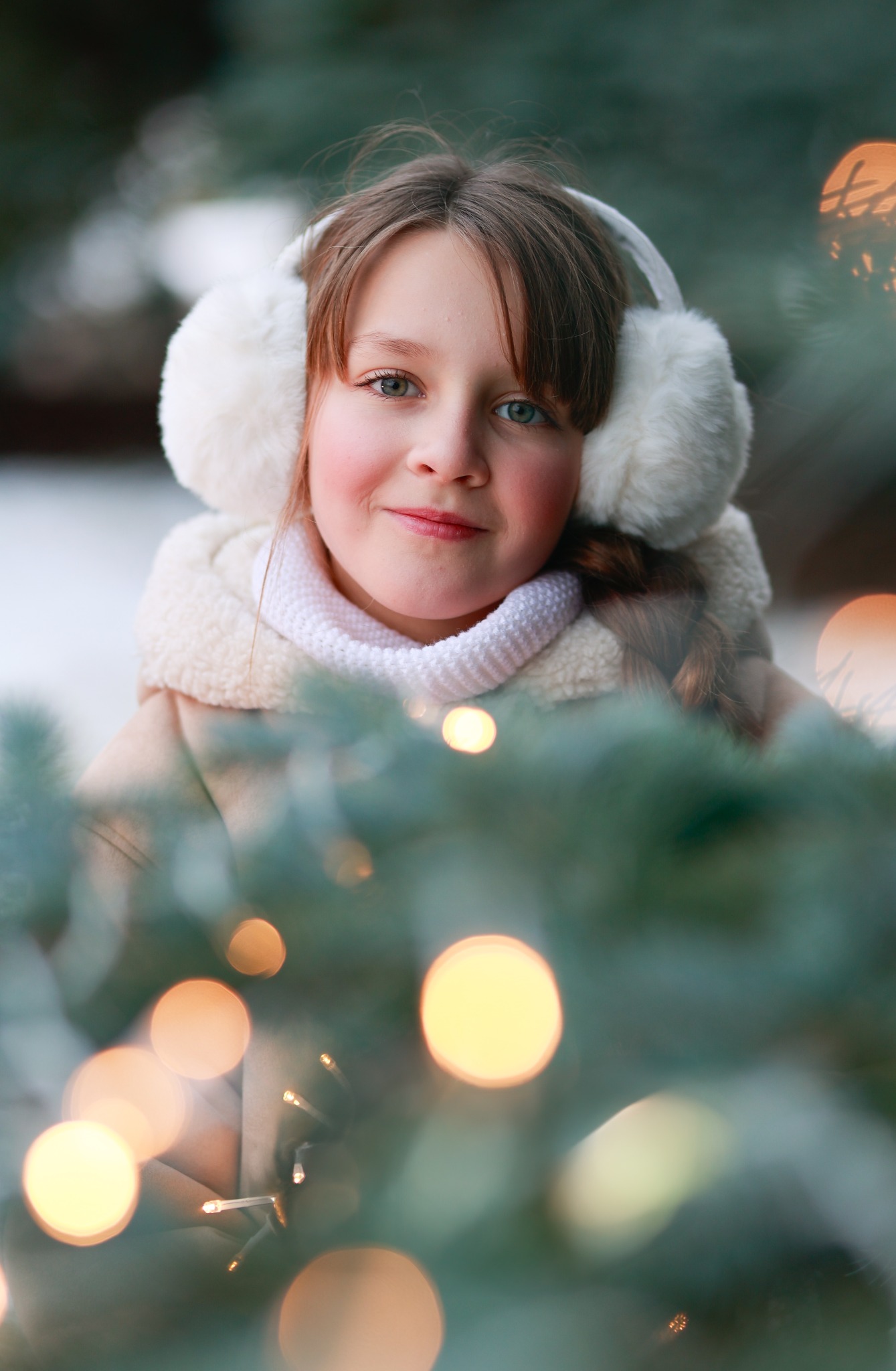 Portret uśmiechniętej dziewczynki w nausznikach, otulonej zimowym szalem, z choinką i światełkami w tle, uchwycony w miękkim, naturalnym świetle.