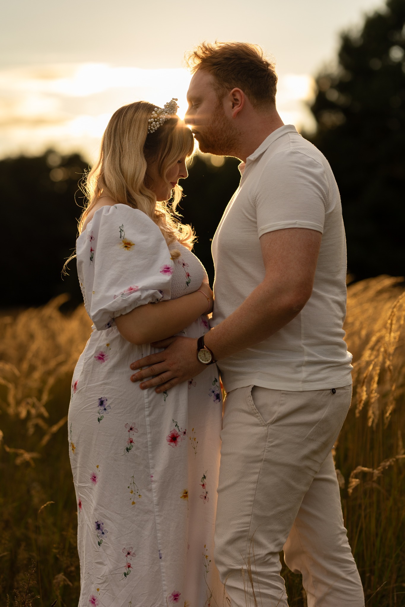 Czułe ujęcie przyszłych rodziców w plenerze o zachodzie słońca. Kobieta w kwiecistej sukience i wianku, mężczyzna w jasnej koszuli, oboje w objęciach, z promieniami słońca.