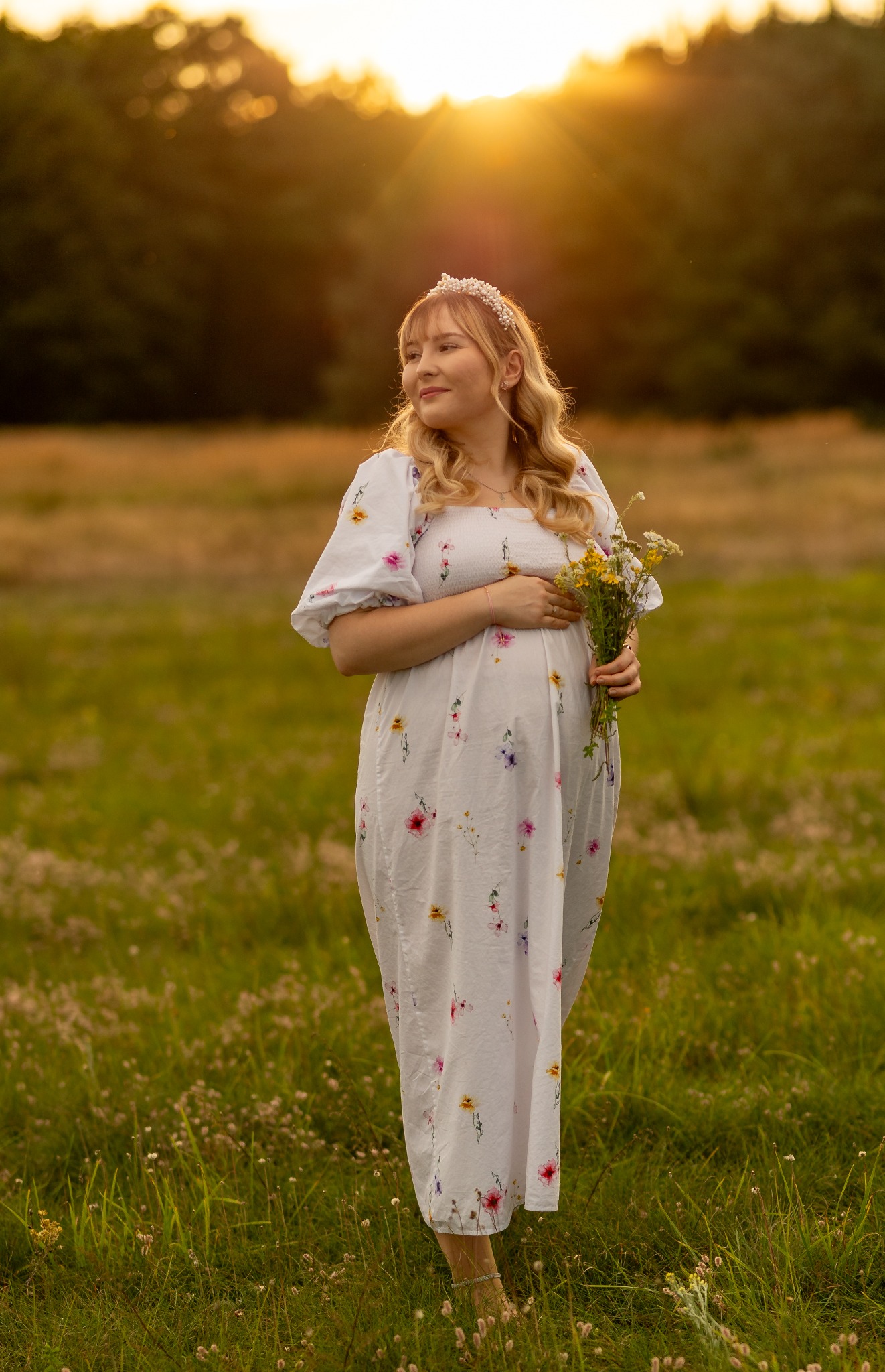 Kobieta w kwiecistej sukience ciążowej, z wiankiem na głowie, trzyma bukiet polnych kwiatów na łące w promieniach zachodzącego słońca. Delikatny uśmiech i wzrok skierowany w bok.