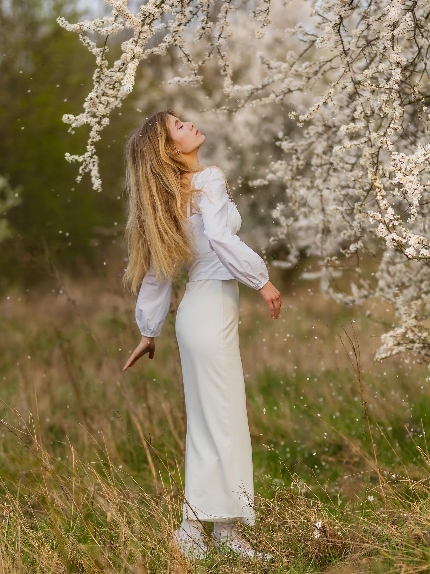 Kobieta w bieli, z zamkniętymi oczami, pod kwitnącym drzewem w plenerze. Delikatne płatki opadają, tworząc romantyczną atmosferę sesji zdjęciowej.